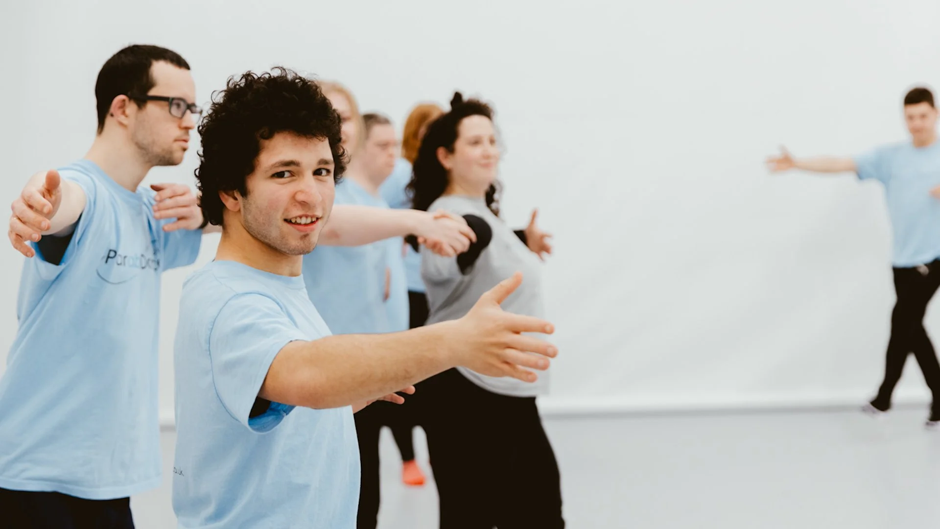 Group of people participating in a dance class in a bright, plain room,  wearing light blue and gray shirts.