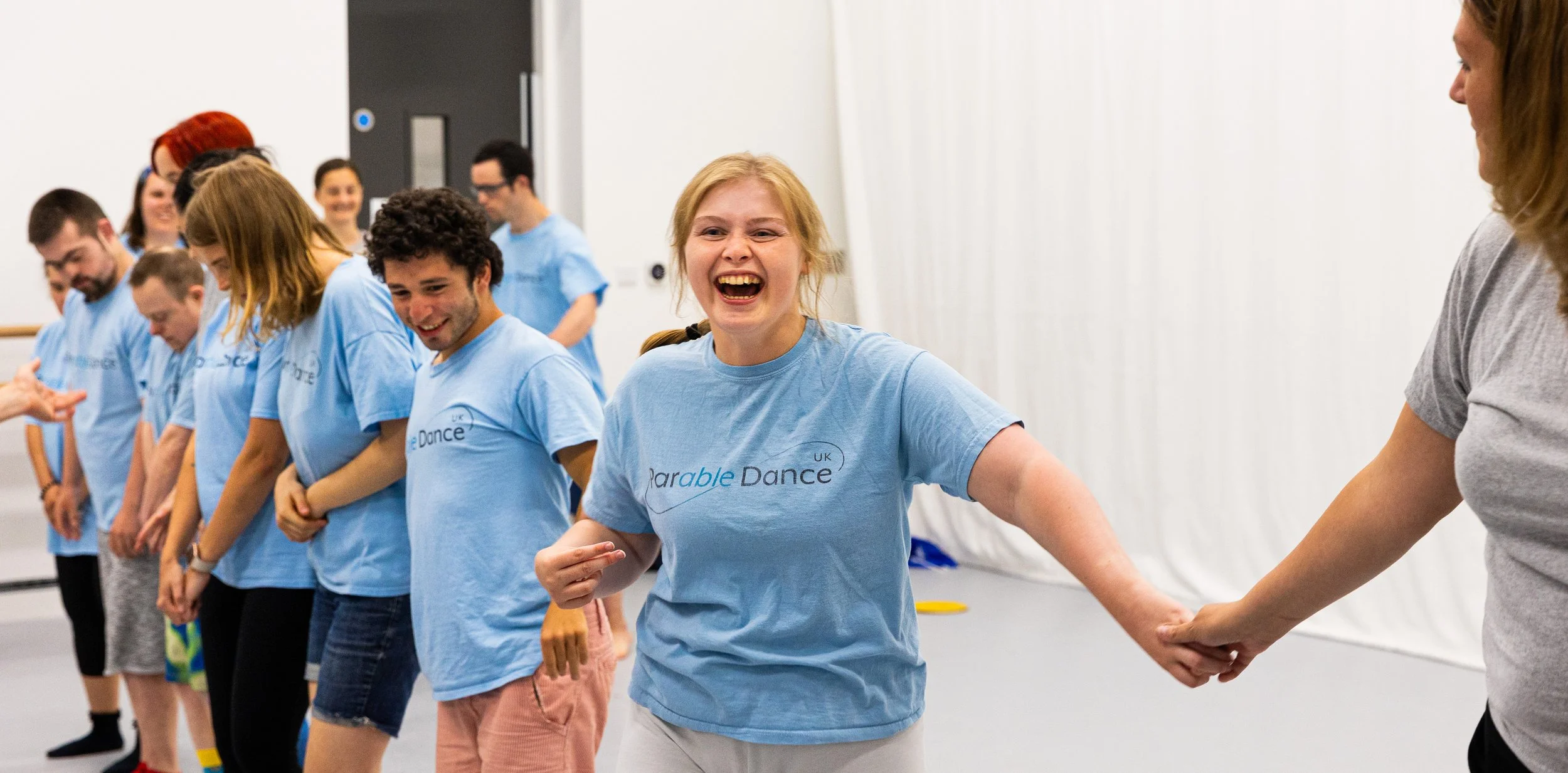 People in a dance class holding hands and laughing, wearing matching light blue shirts, in a studio with a white curtain backdrop.