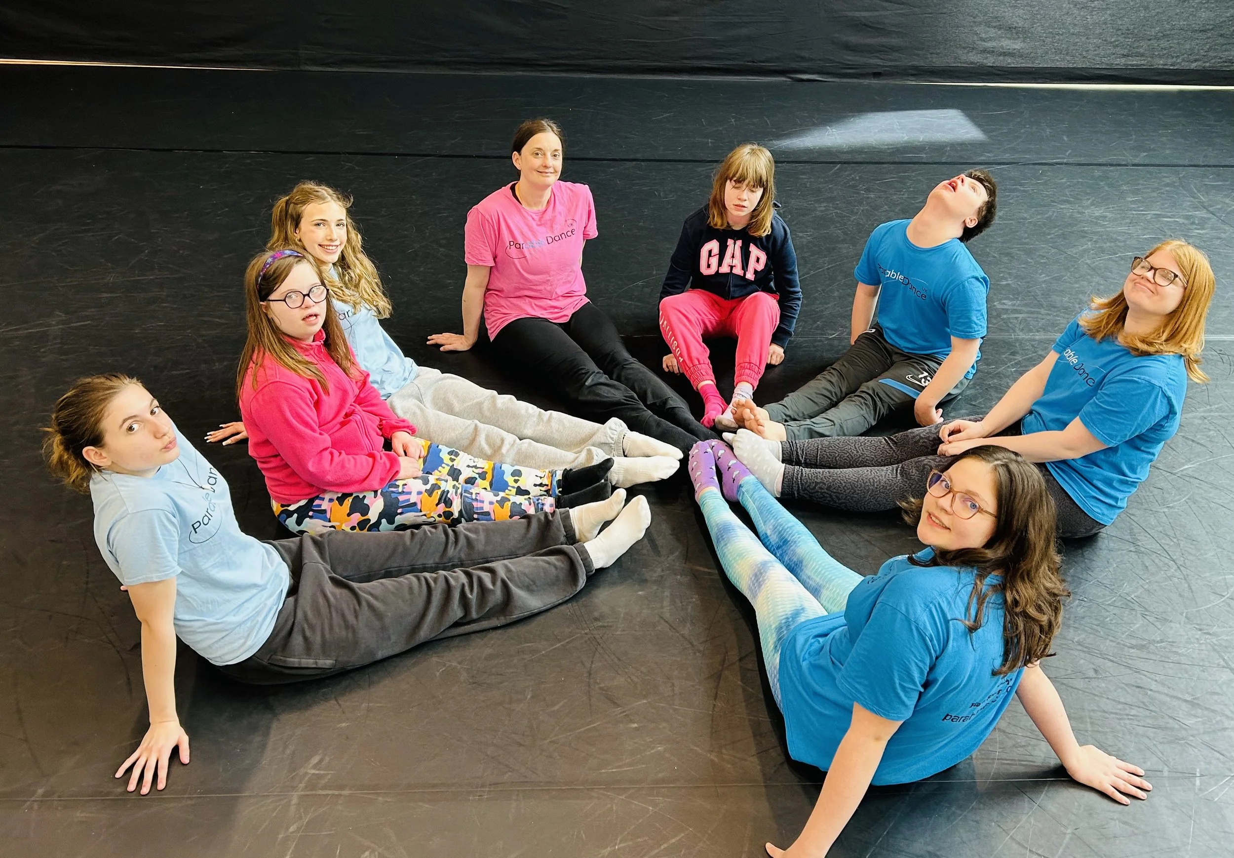 A group of nine girls and a woman sitting on a black dance studio floor, forming a circle with their legs extended toward each other, smiling and looking at the camera.