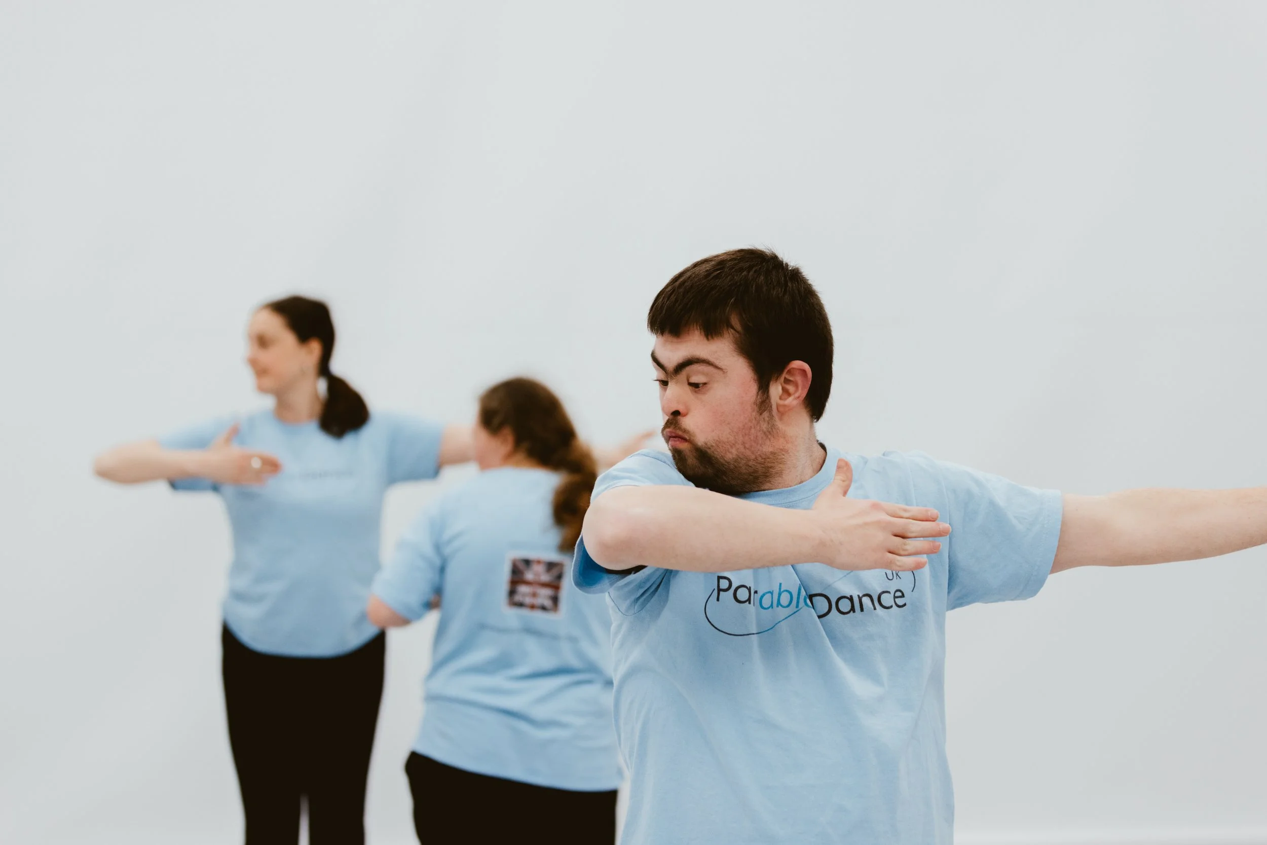 People participating in a dance class, performing stretching or warm-up exercises, wearing light blue shirts with the Parable Dance logo.