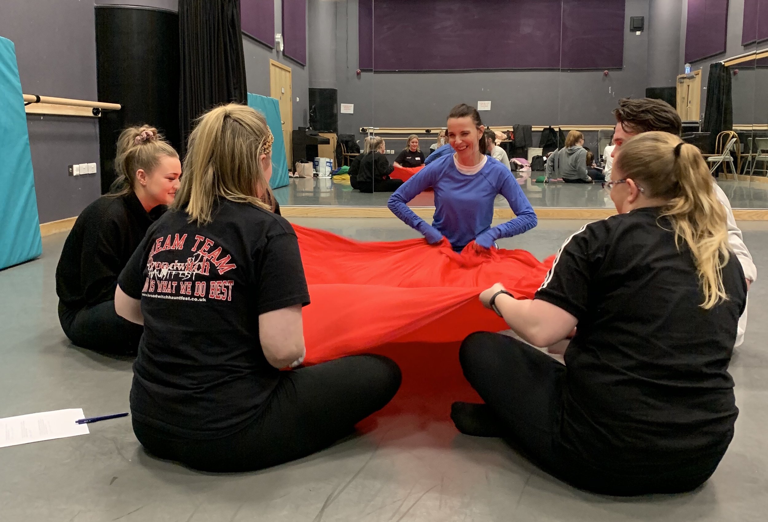 Group of women sitting on the floor in a dance studio, holding a red parachute.