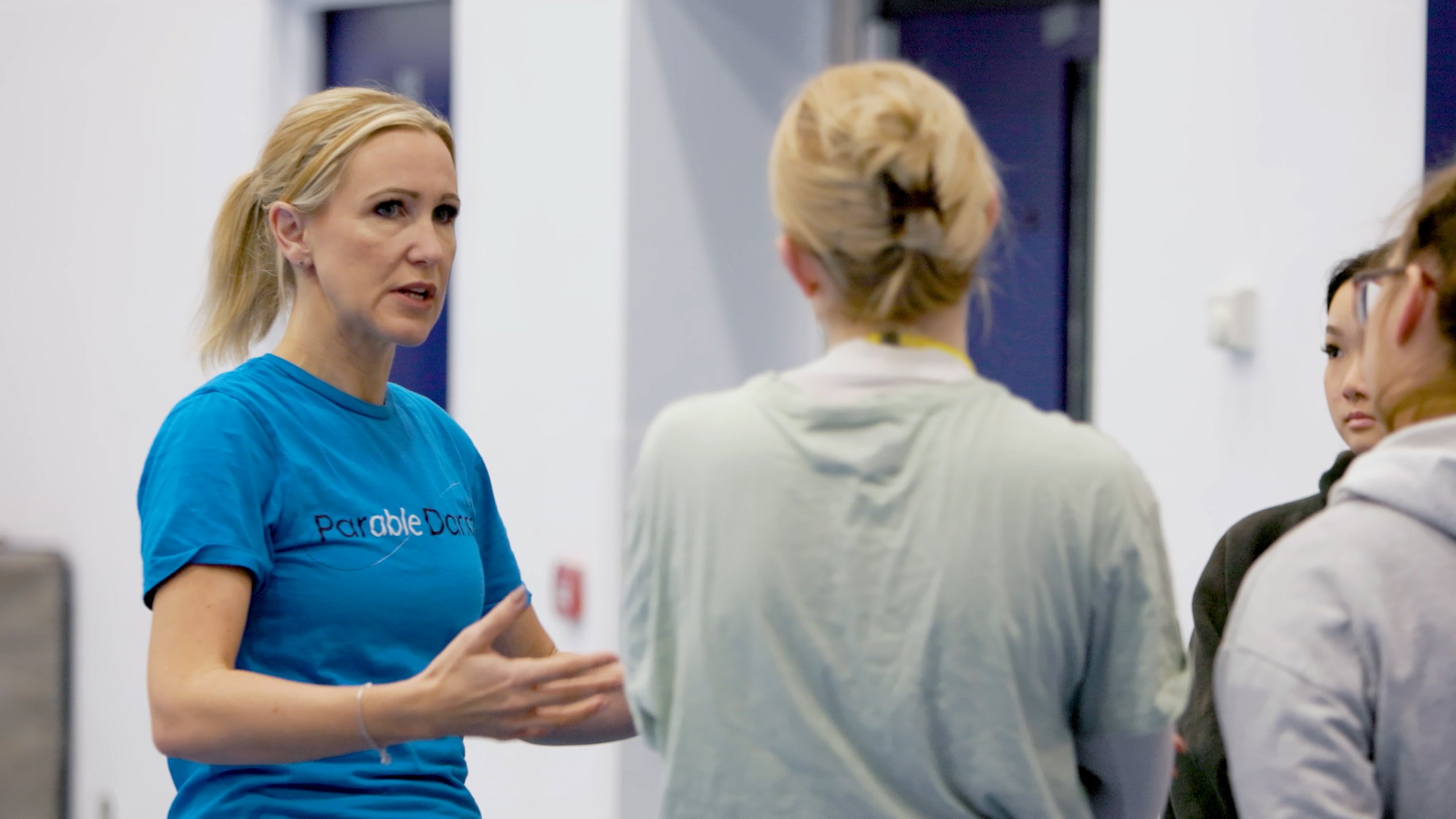 A woman with blonde hair wearing a blue shirt with 'ParableDance' written on it is speaking to three other women. She appears to be explaining something in a room with white walls and a dark blue door.
