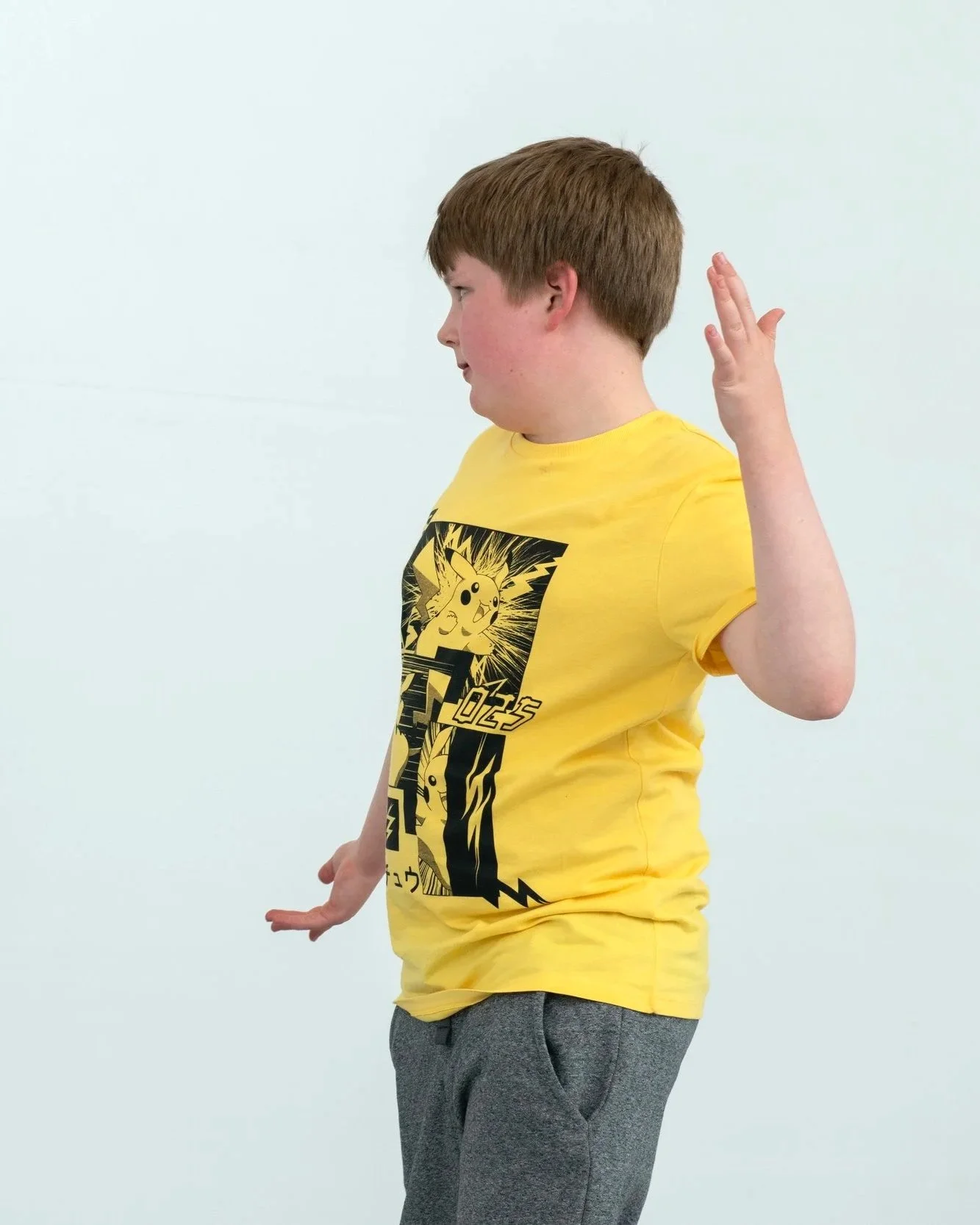 A young boy with brown hair wearing a yellow 'Pokemon' t-shirt and gray trousers standing against a plain white background, holding his right hand up with fingers extended.