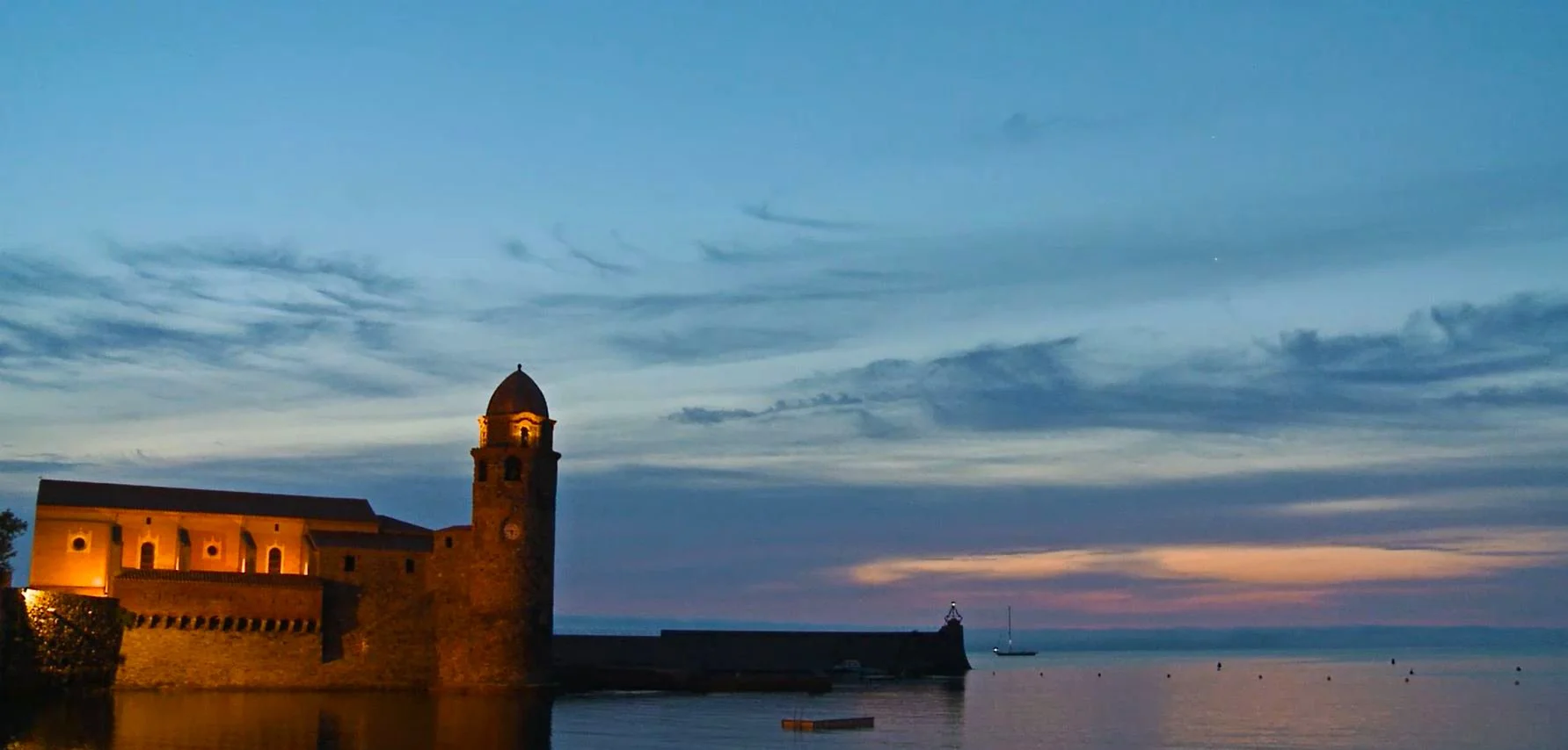 Coucher de soleil sur un bâtiment ancien en pierre situé au bord d'une mer calme, avec une voile au loin dans le ciel bleu