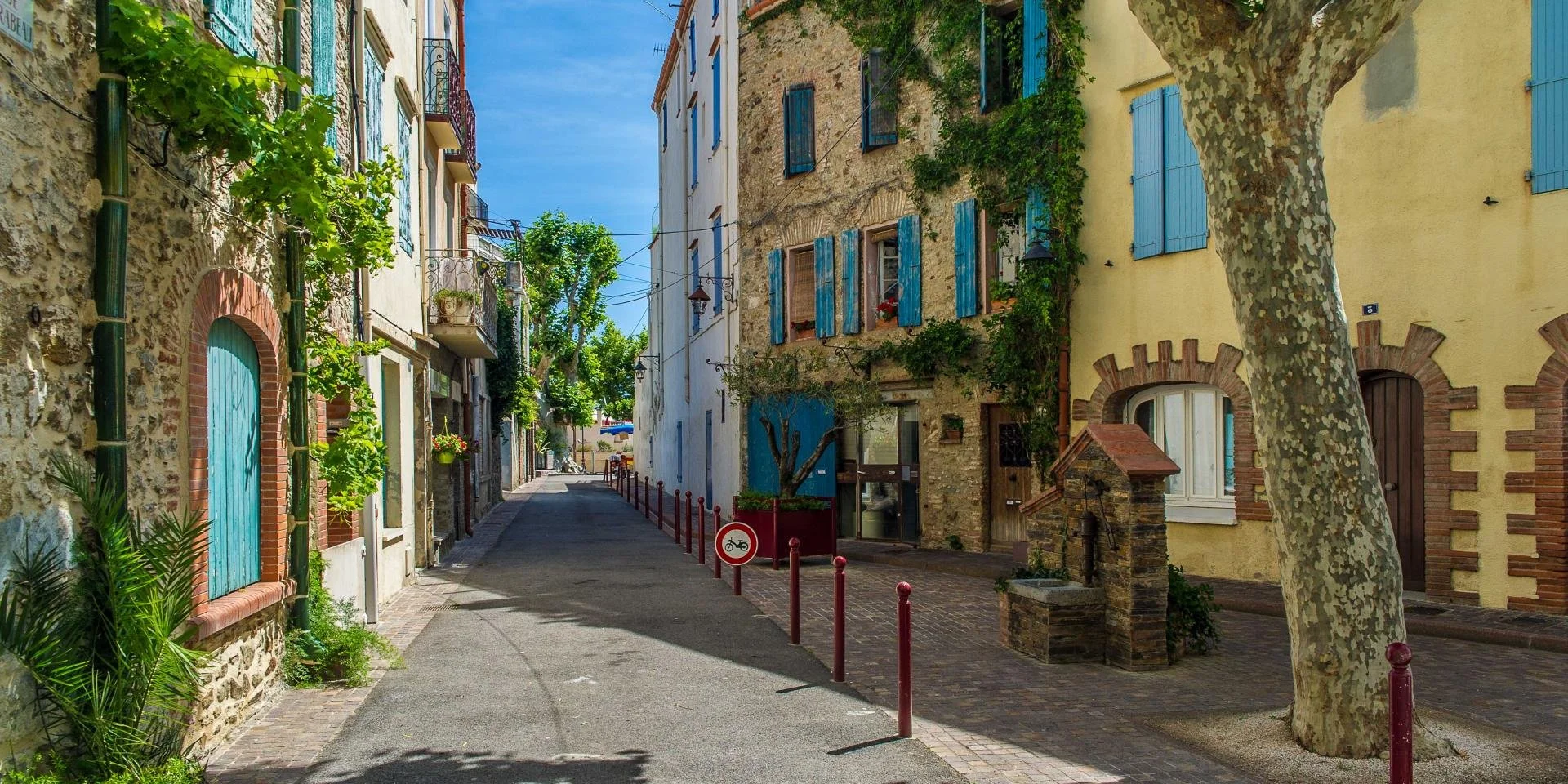 Rue pavée de Banyuls sur mer bordée de maisons colorées avec volets bleus, arbres, barrières rouges et panneau pour vélo en ville ensoleillée.