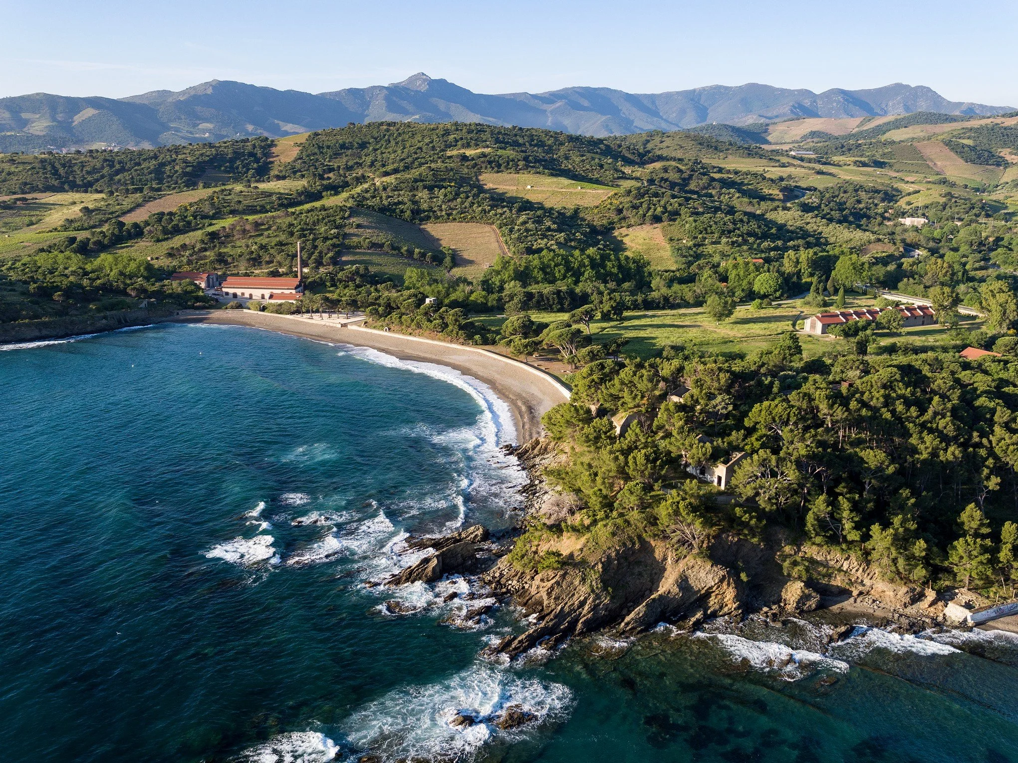 Vue aérienne d'une côte rocheuse, la cote vermeille, avec une plage de sable et des vagues, entourée de collines verdoyantes et de montagnes en arrière-plan.