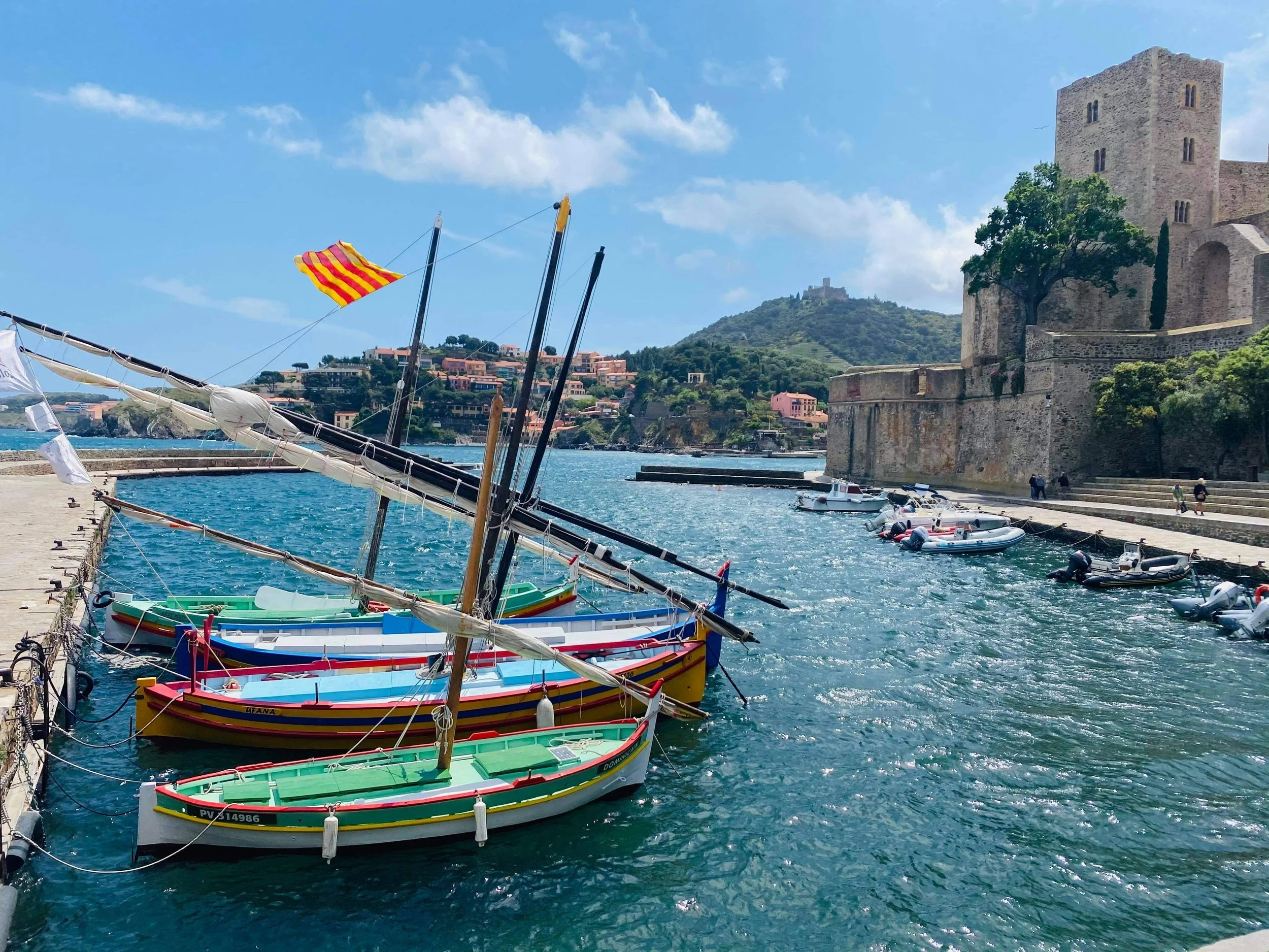 Petits bateaux Catalans colorés amarrés dans un port, avec un château en pierre et une colline verdoyante en arrière-plan, sous un ciel ensoleillé avec quelques nuages.
