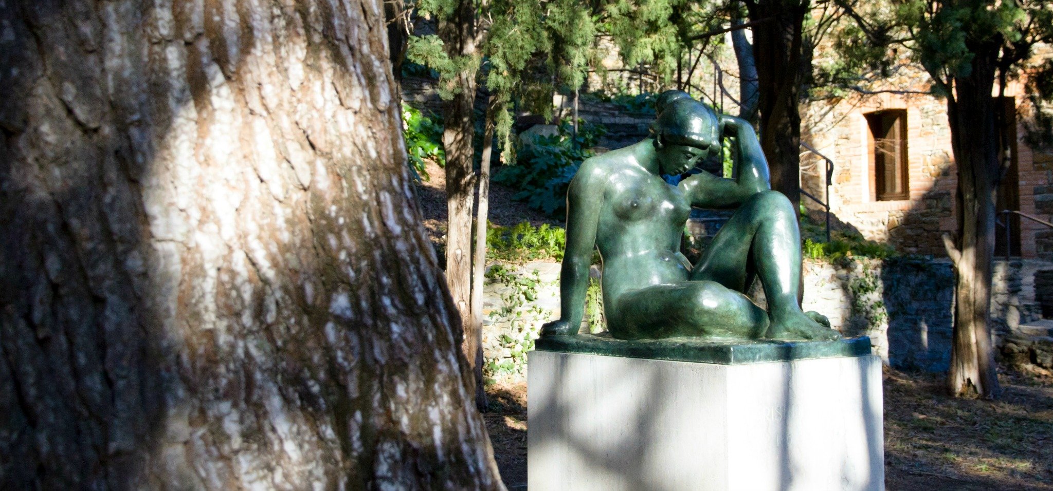 Sculpture de Maillol d'une femme nue assise dans un jardin avec des arbres et une maison en arrière-plan.