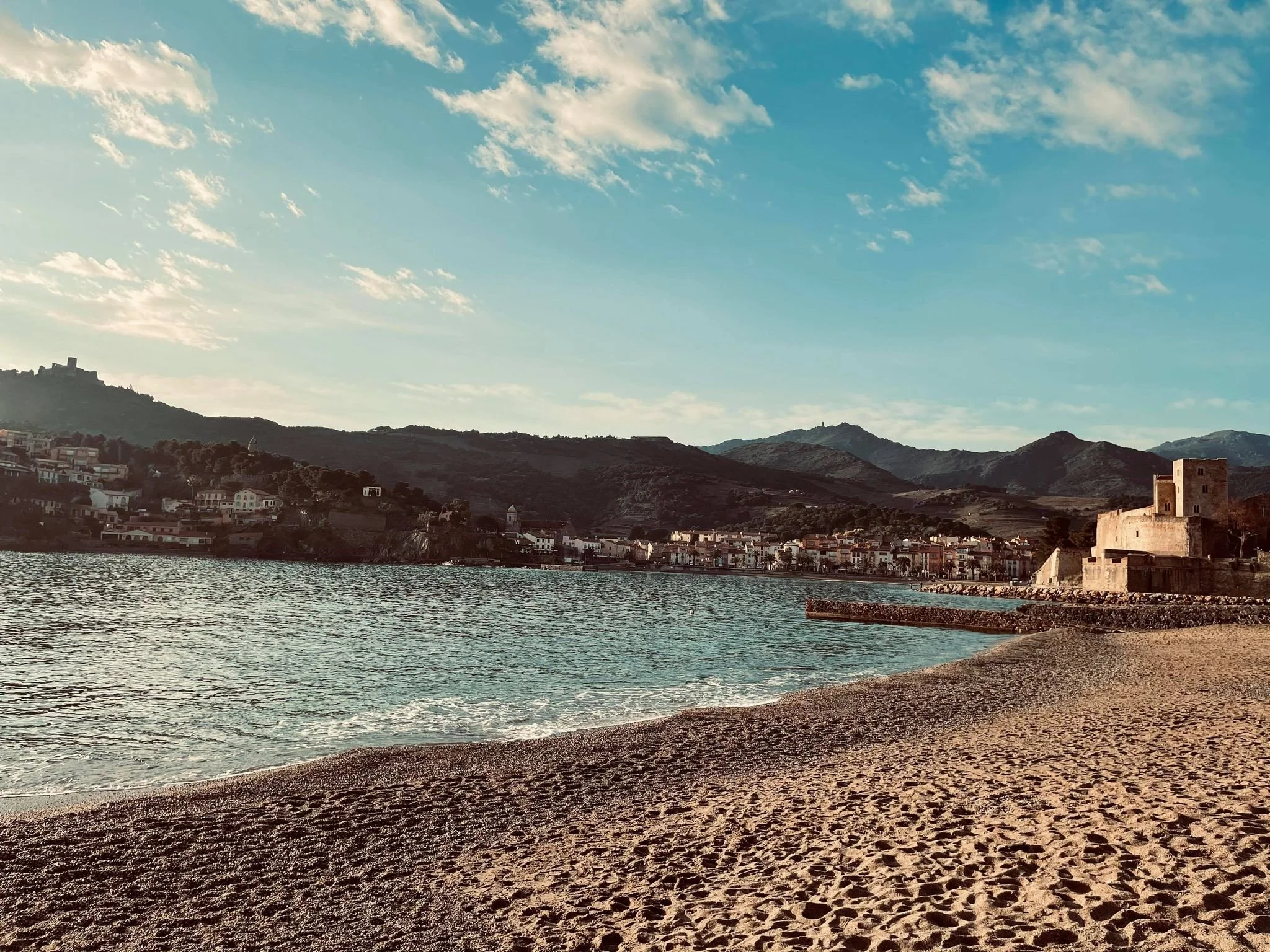 Plage de sable Collioure avec des vagues, ville au fond avec des maisons, montagnes derrière, et un ciel bleu avec quelques nuages.