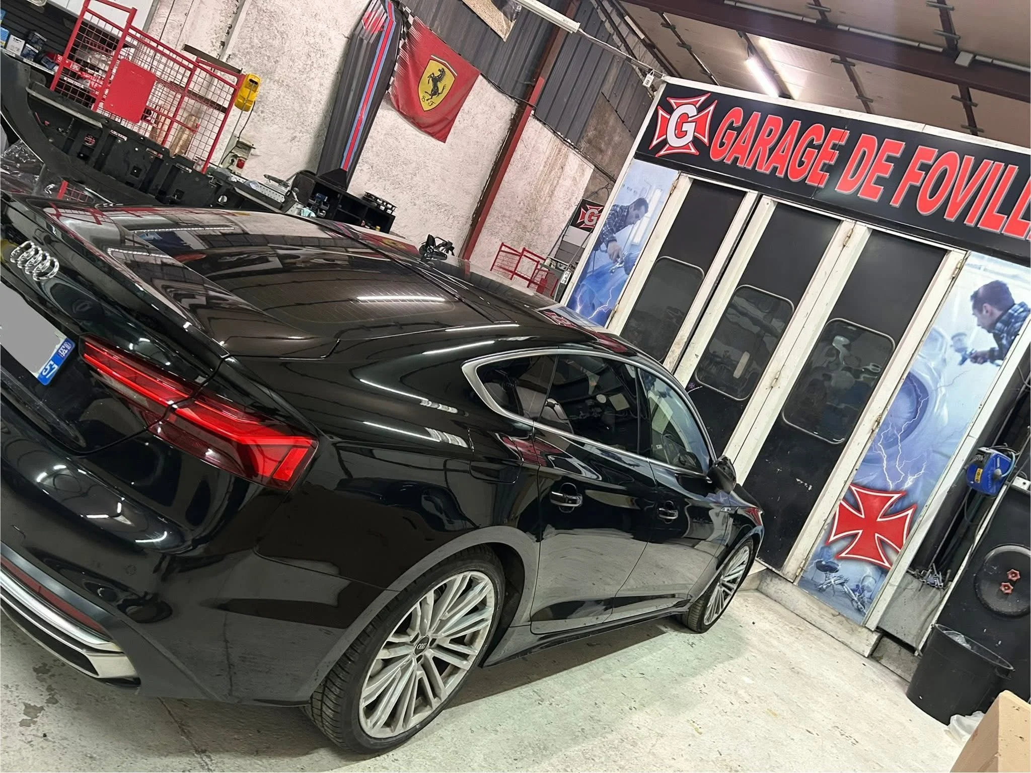 Voiture noire dans un garage avec une bannière et une affiche de Ferrari, logo de la marque automobile Audi visible sur la voiture.