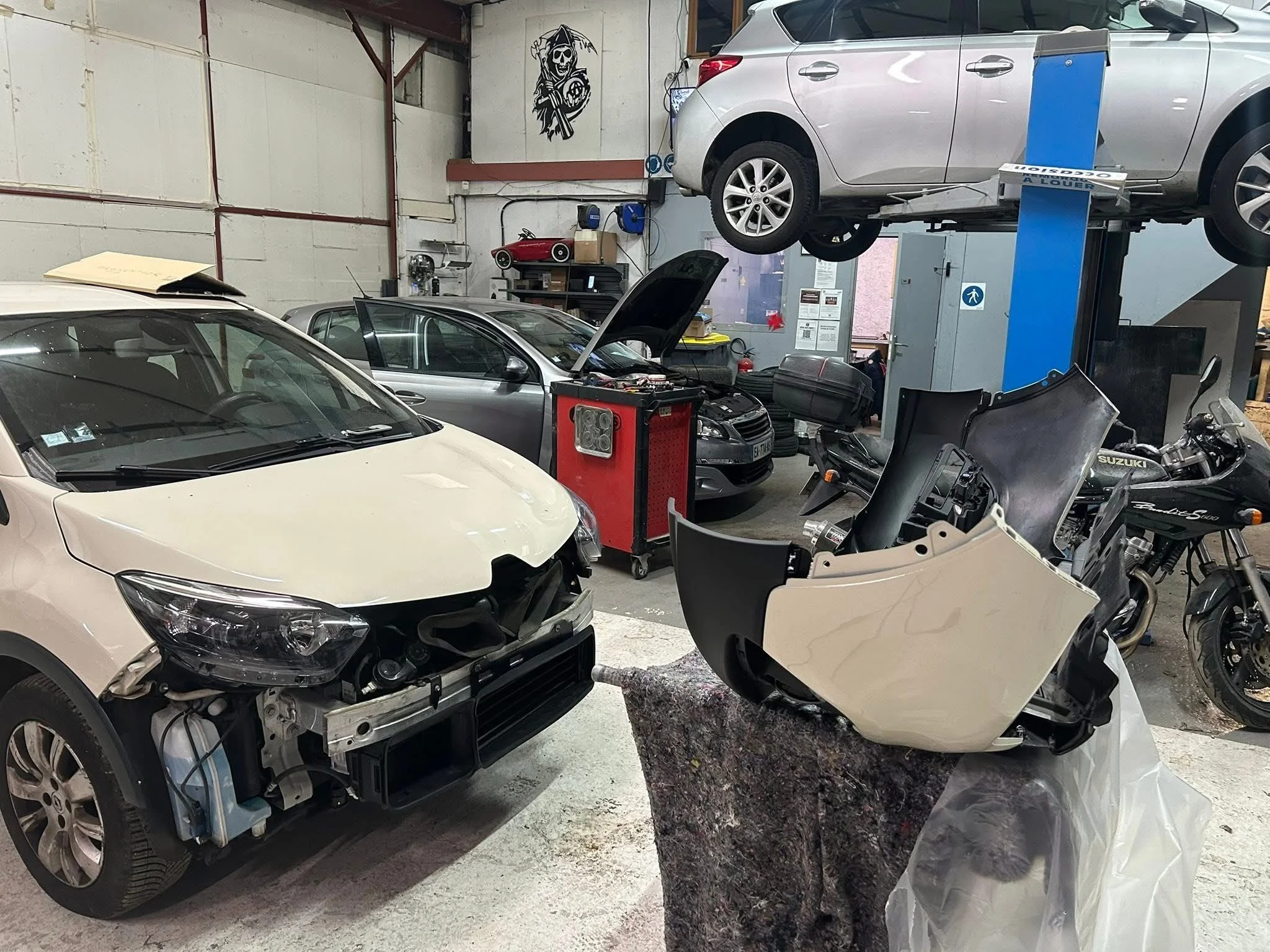 Un garage ou atelier de réparation automobile avec plusieurs voitures, dont une en réparation avec le capot ouvert et une autre sur un pont élévateur. Il y a aussi des pièces de voiture, un moteur et des motos dans l'atelier.