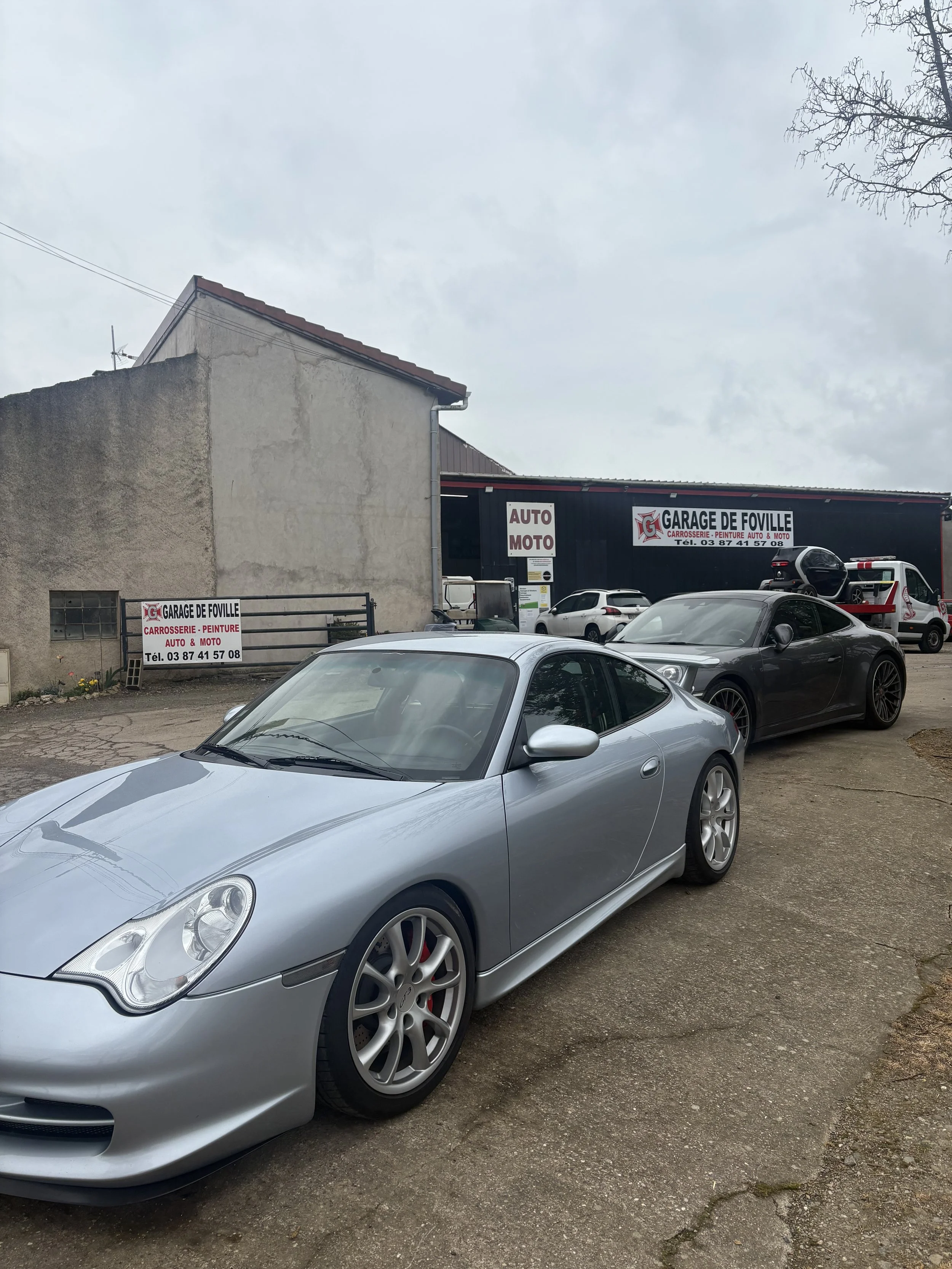 voiture de sport argentée garée devant un atelier de réparation auto nommé Garage de Foville, avec d'autres voitures en arrière-plan, bâtiment en arrière-plan, ciel nuageux