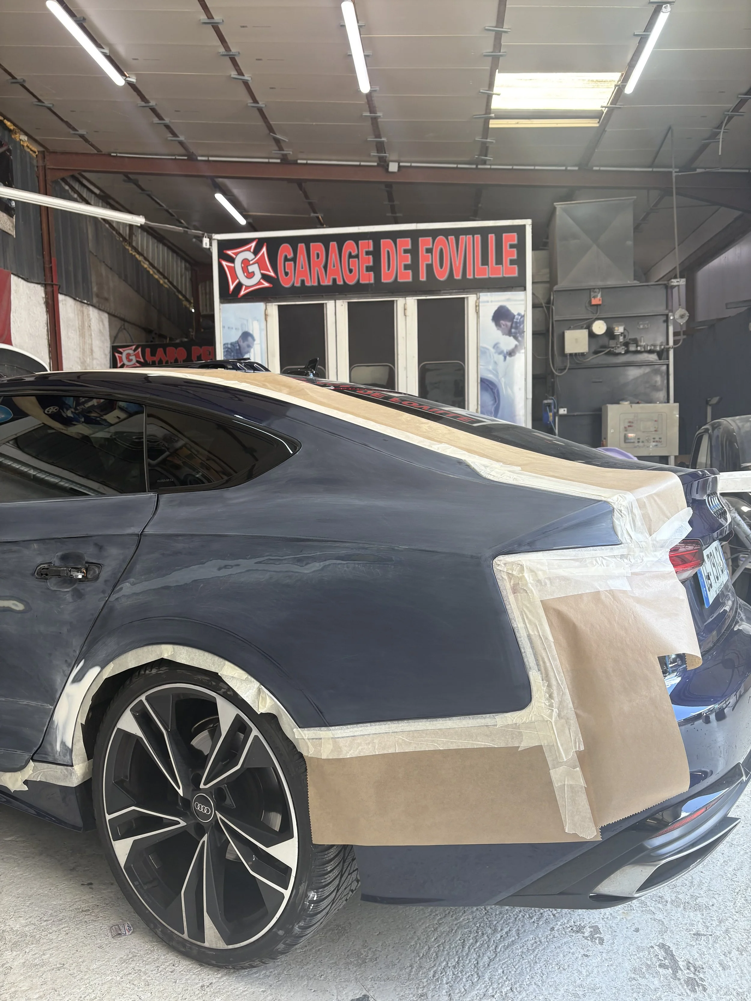 Voiture noire en réparation dans un garage, avec du ruban de masquage et du papier pour la peinture, devant un panneau indiquant 'Garage de Foville'.
