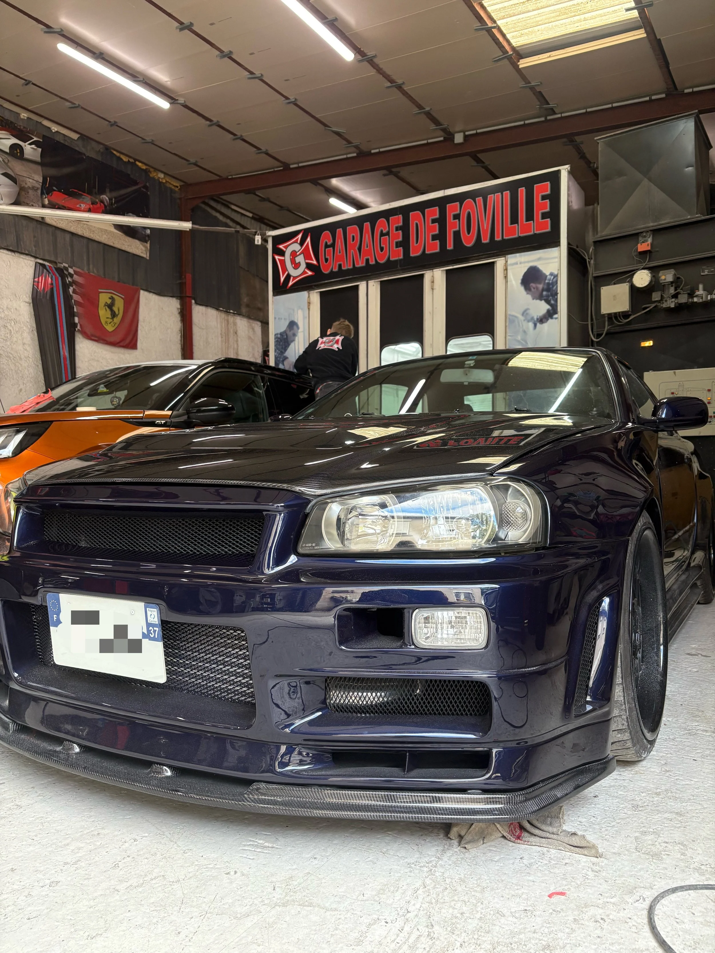 Voiture noire de sport dans un garage, avec d'autres voitures visibles en arrière-plan, et un panneau indiquant « Garage de Foville ».