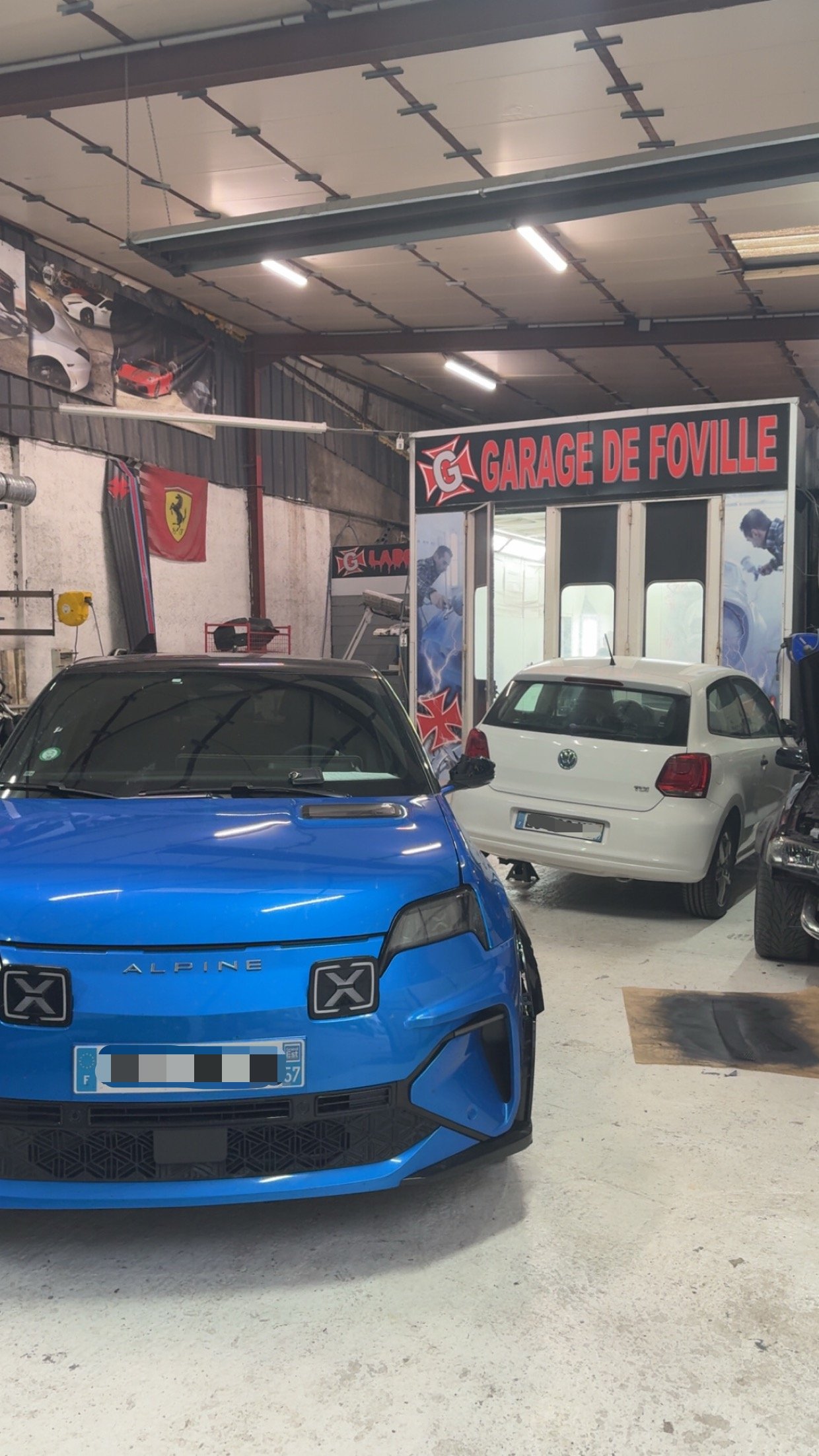 Intérieur d'un garage automobile avec plusieurs voitures, y compris une voiture bleue Alpine en premier plan et une voiture blanche en arrière-plan, avec une enseigne 'Garage de Foville' et des affiches de voitures sur le mur.