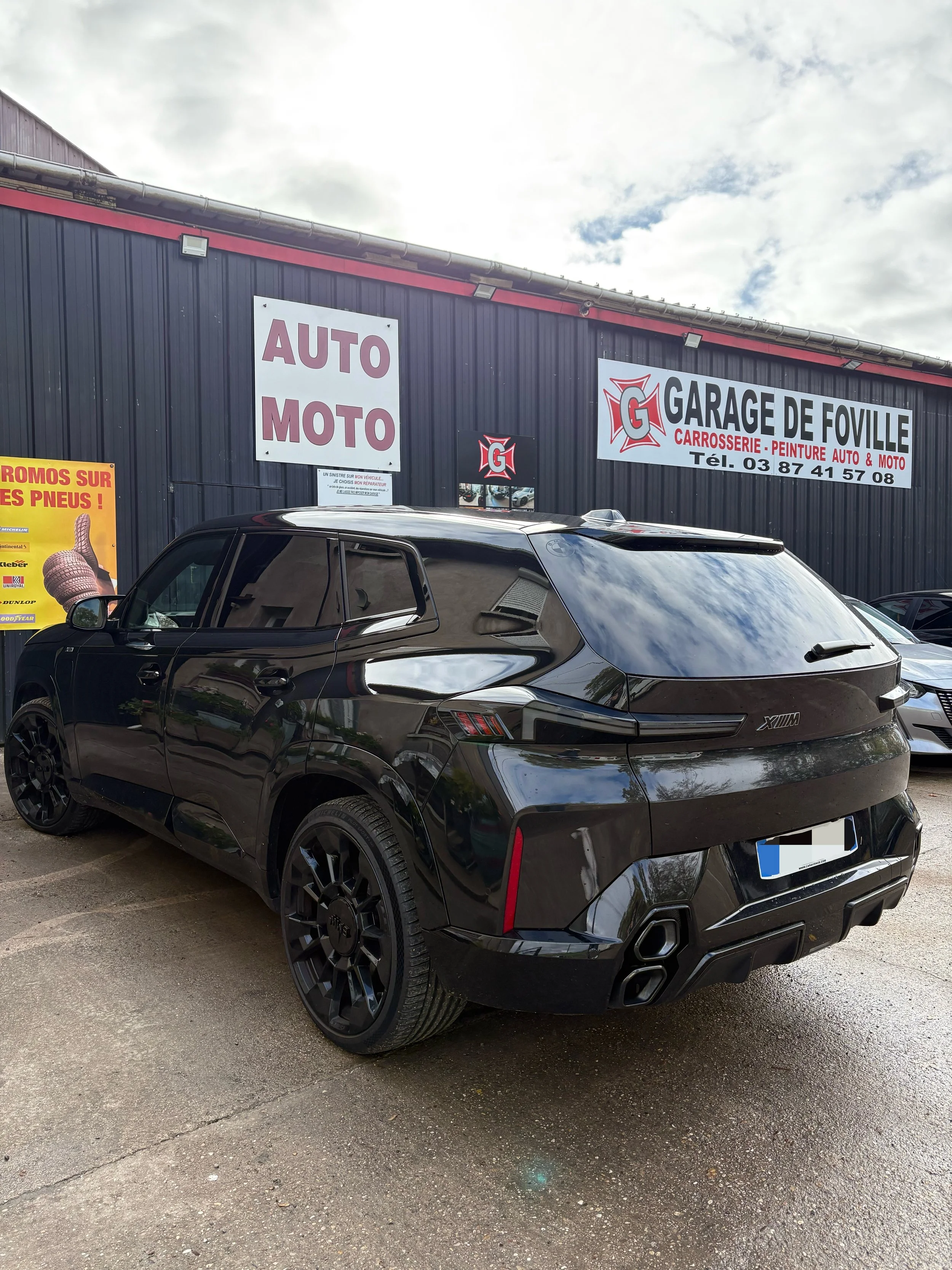 Voiture noire SUV garée devant un garage de réparation auto, avec panneaux publicitaires et un bâtiment gris en arrière-plan.