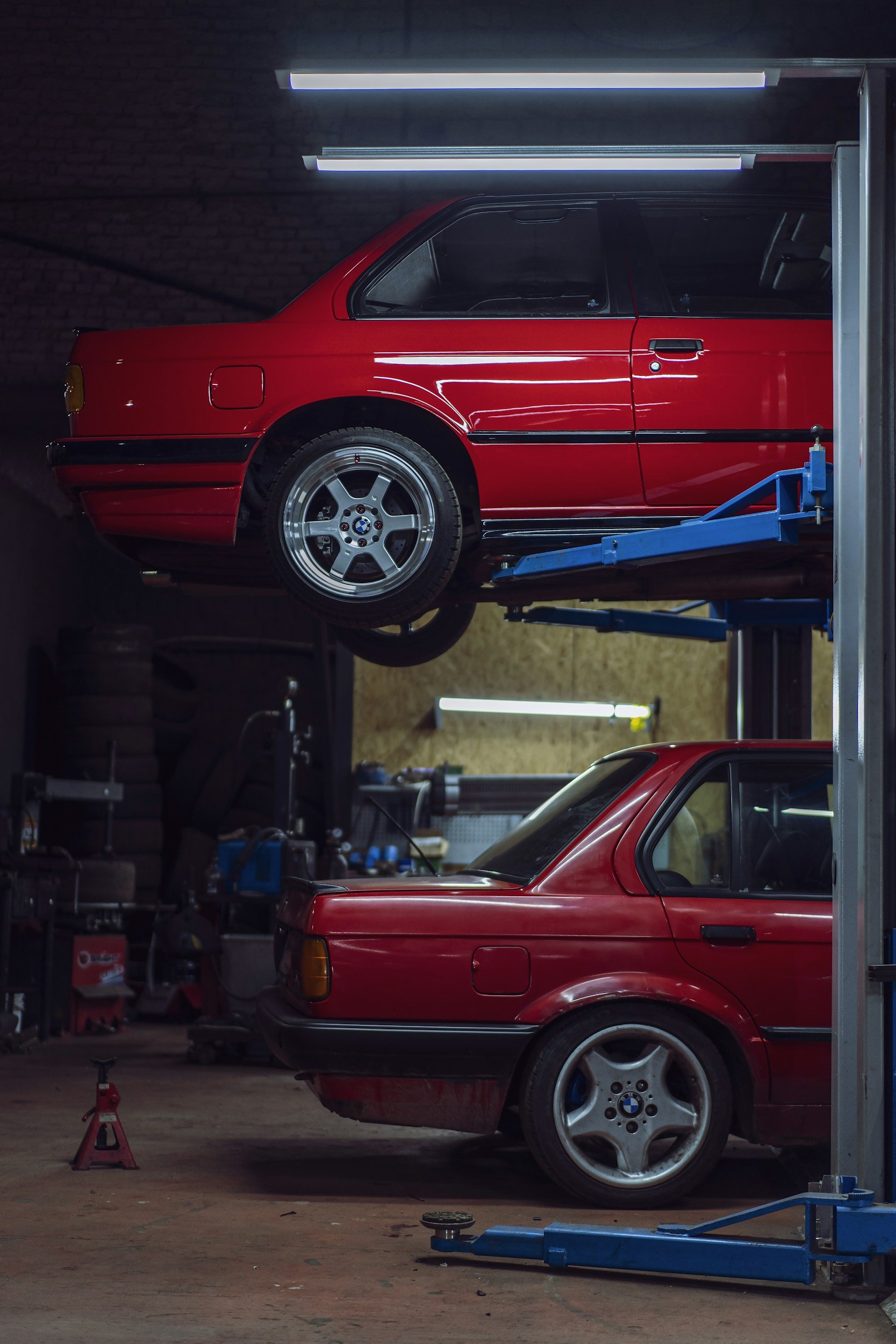 Two red BMW cars in a garage, one on a lift and the other on the ground, with various tools and equipment around.