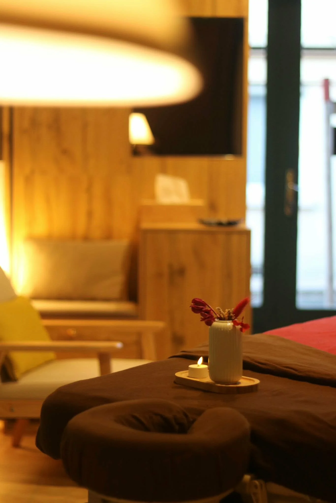 Intérieur d'une chambre avec une bougie allumée et un vase avec des fleurs sur le lit en premier plan, meubles en bois au fond, lumière tamisée.