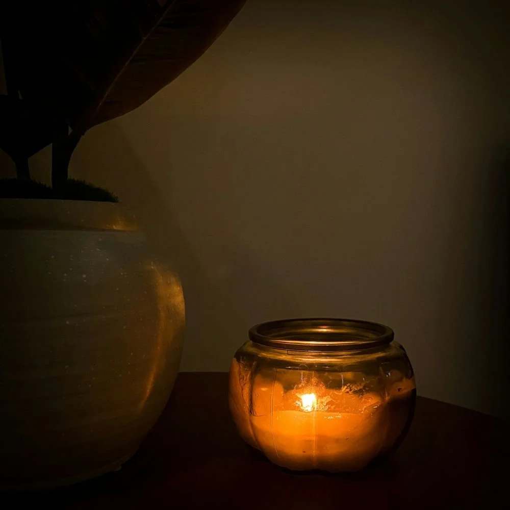 Une bougie allumée dans un pot en verre posé sur une surface en bois, avec un grand vase à côté, dans une ambiance tamisée.
