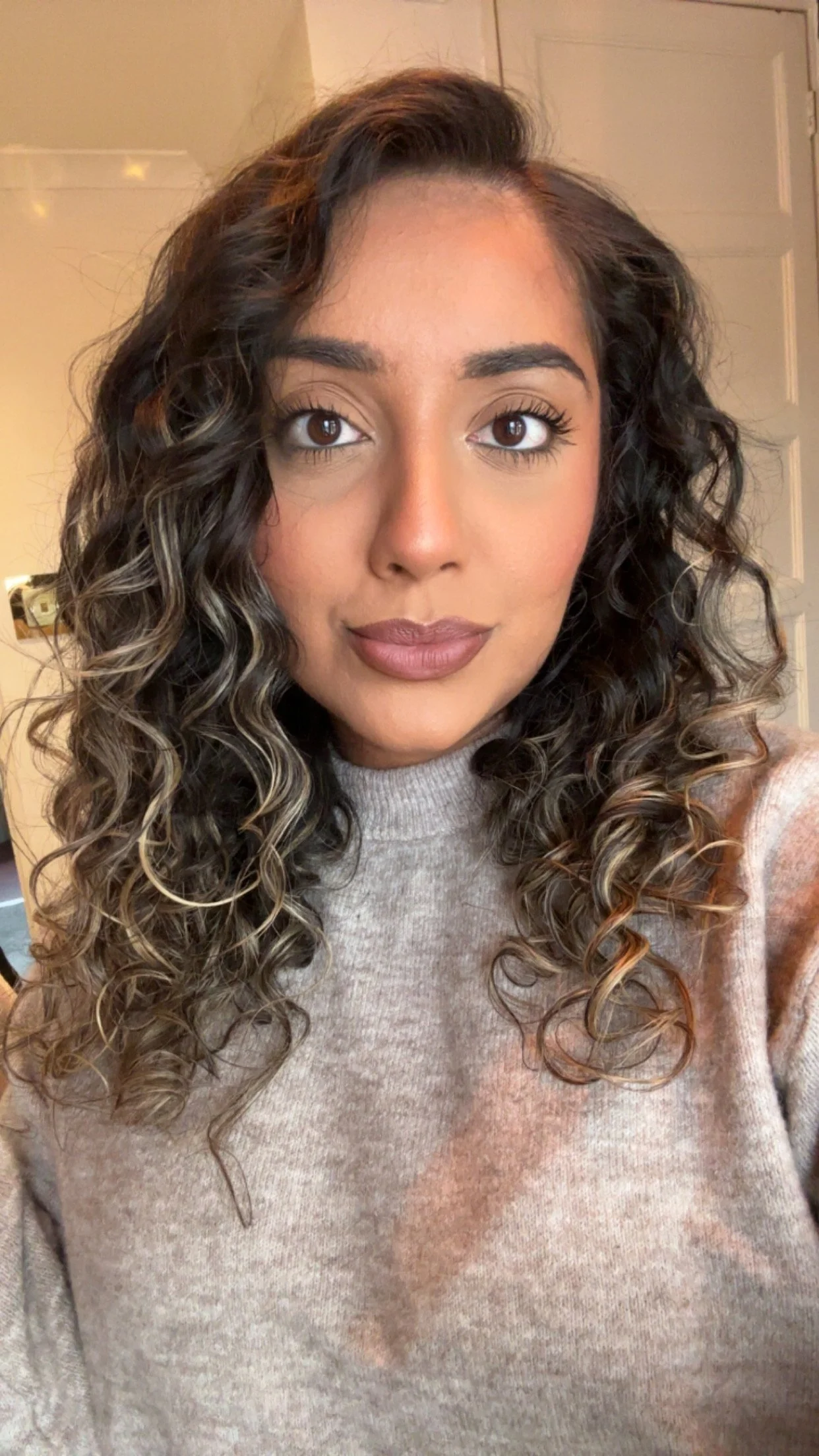 A woman with curly brown and blonde hair taking a selfie indoors, wearing a beige sweater.