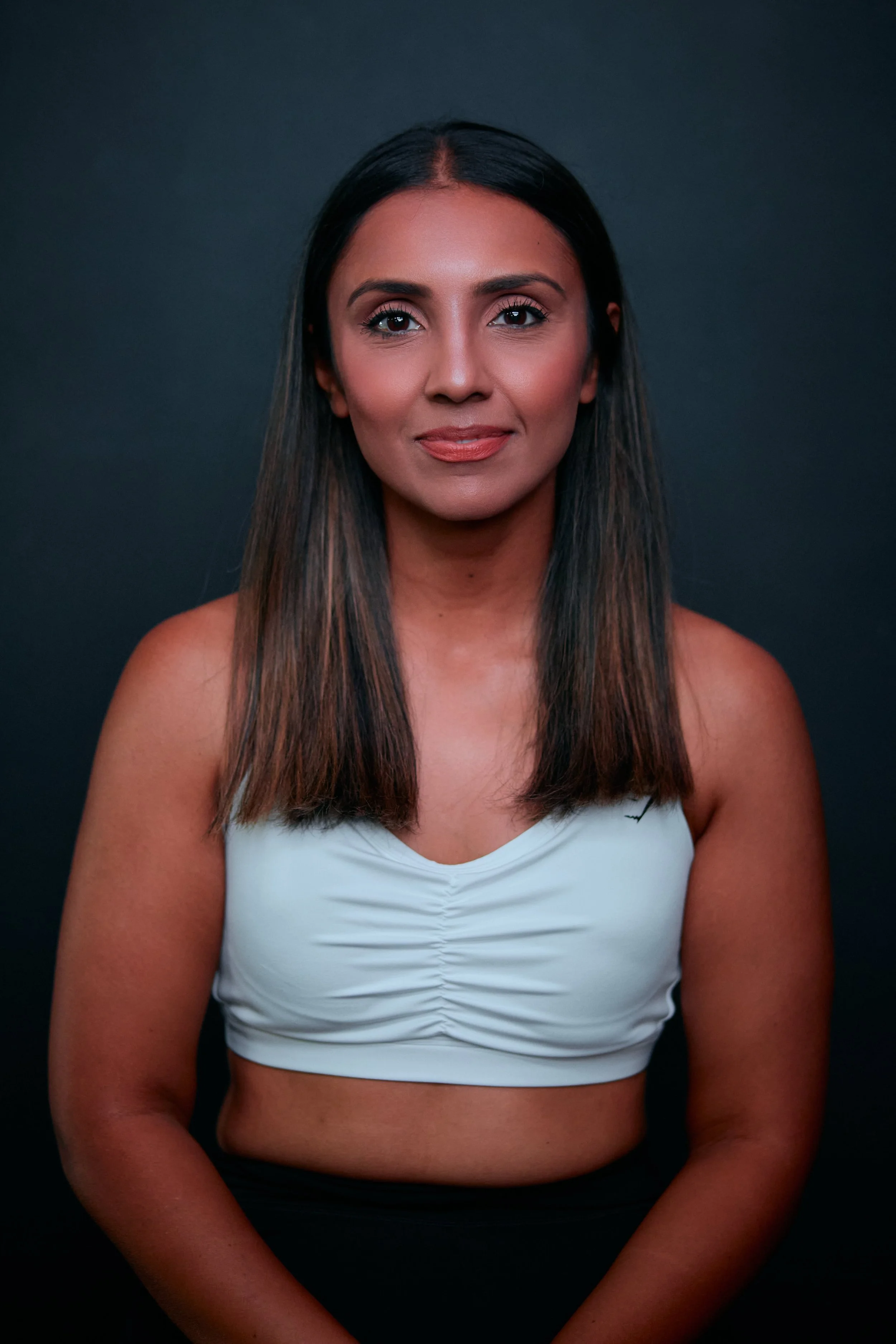 A woman with long dark hair, wearing a white athletic top, standing against a dark background, looking at the camera with a slight smile.
