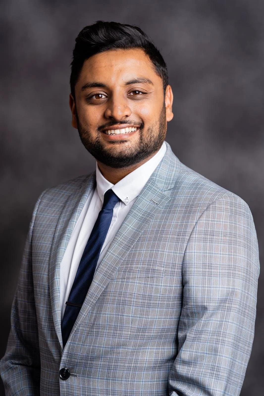 A young man with dark hair and a beard wearing a light gray checkered suit, white shirt, and navy tie, smiling against a dark gray background.
