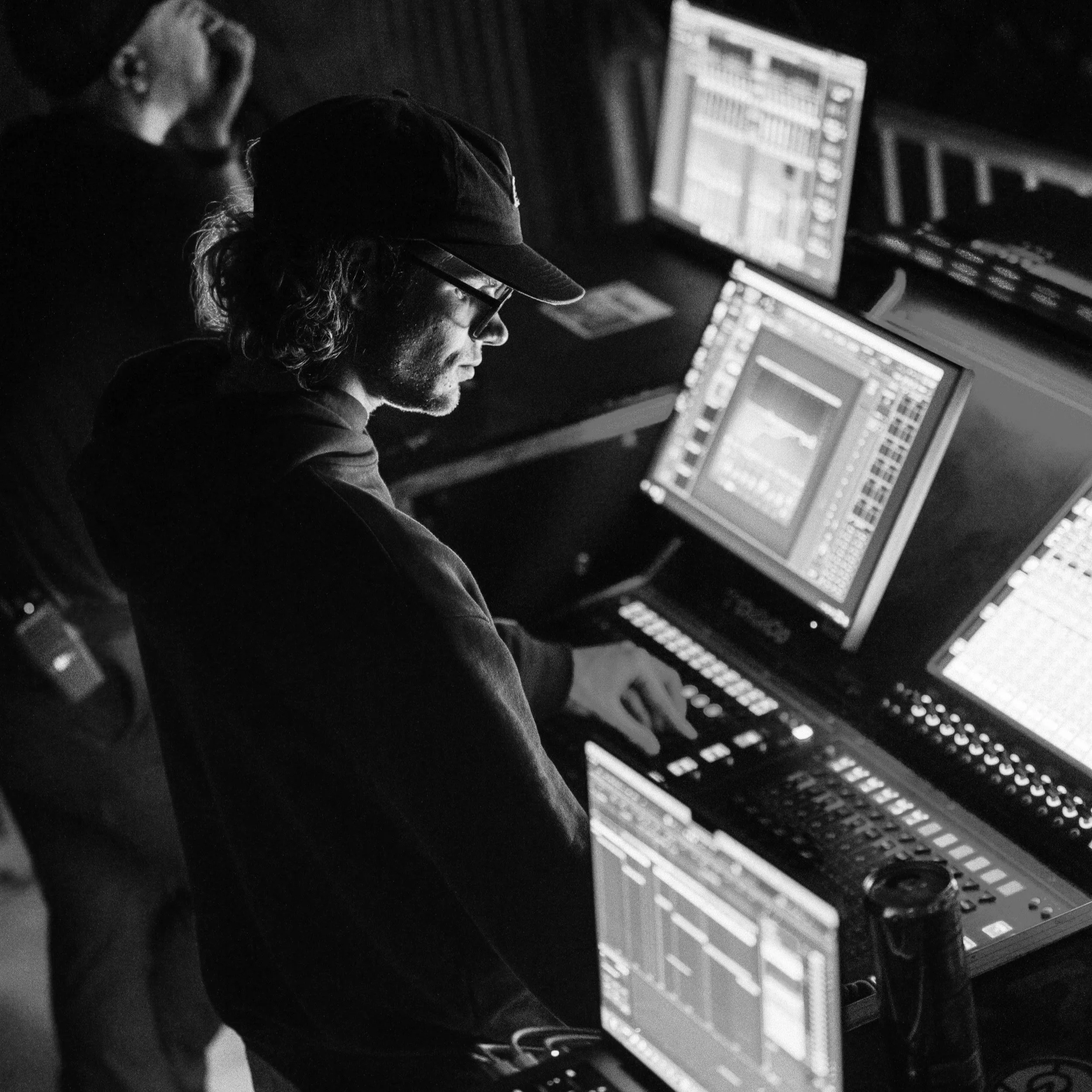 A person with shoulder-length hair, wearing a cap and glasses, working on a music production console with multiple screens displaying music software.