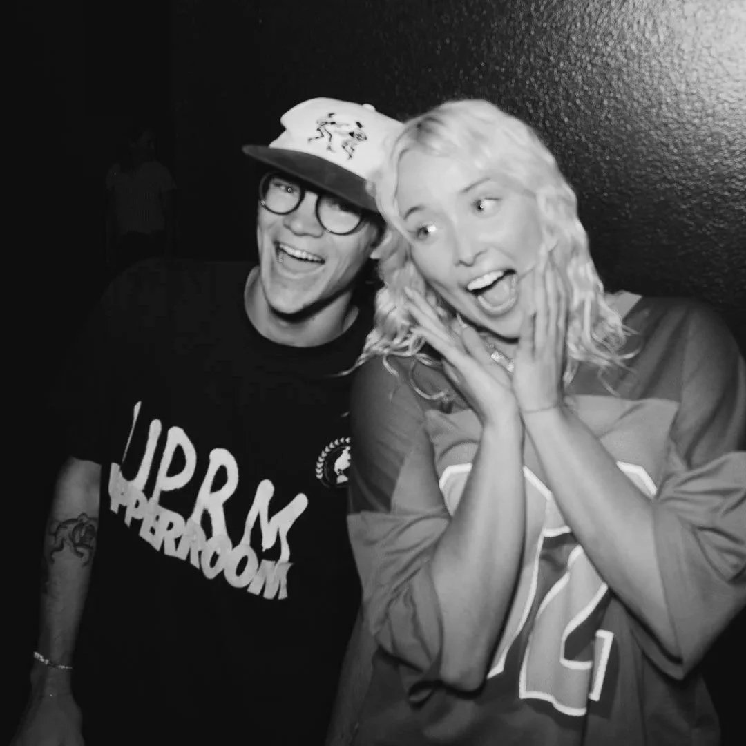 Two friends smiling and posing together in a black and white photo, one wearing glasses and a cap, the other with curly hair and a sports jersey.