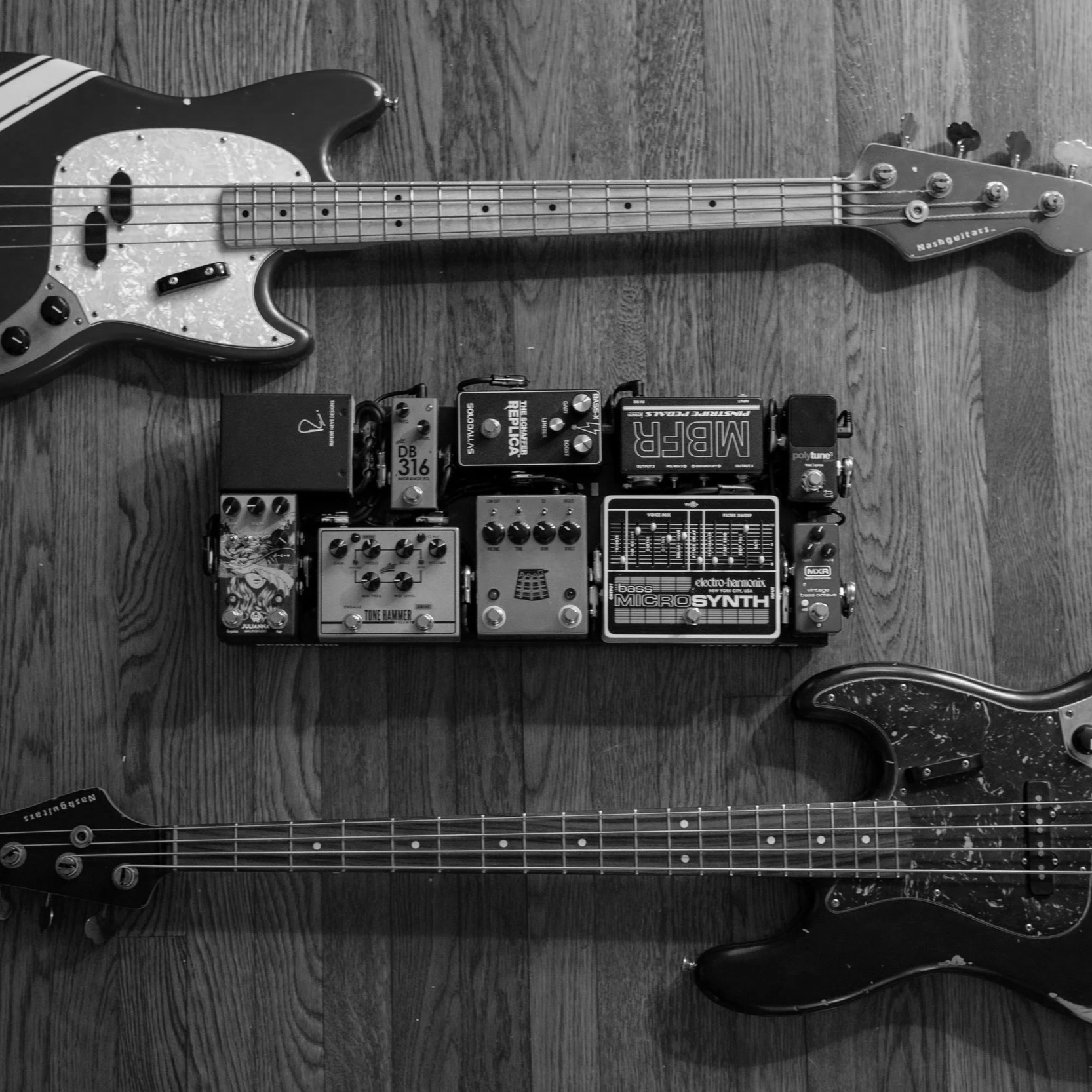 Black and white photo of two electric bass guitars head-to-head with a pedalboard in between on a wooden floor.