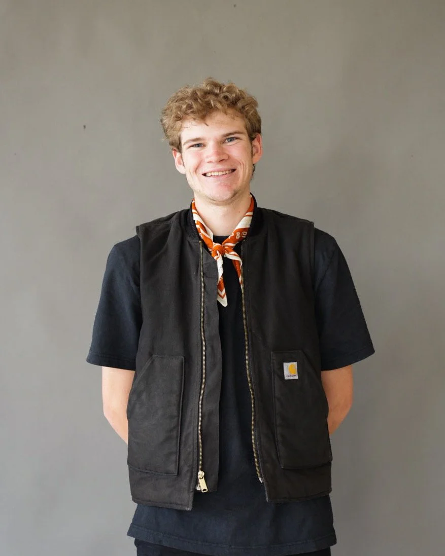 Young man with curly blond hair smiling, wearing a black Carhartt vest, black t-shirt, and a brown and orange neckerchief, standing against a plain gray background.