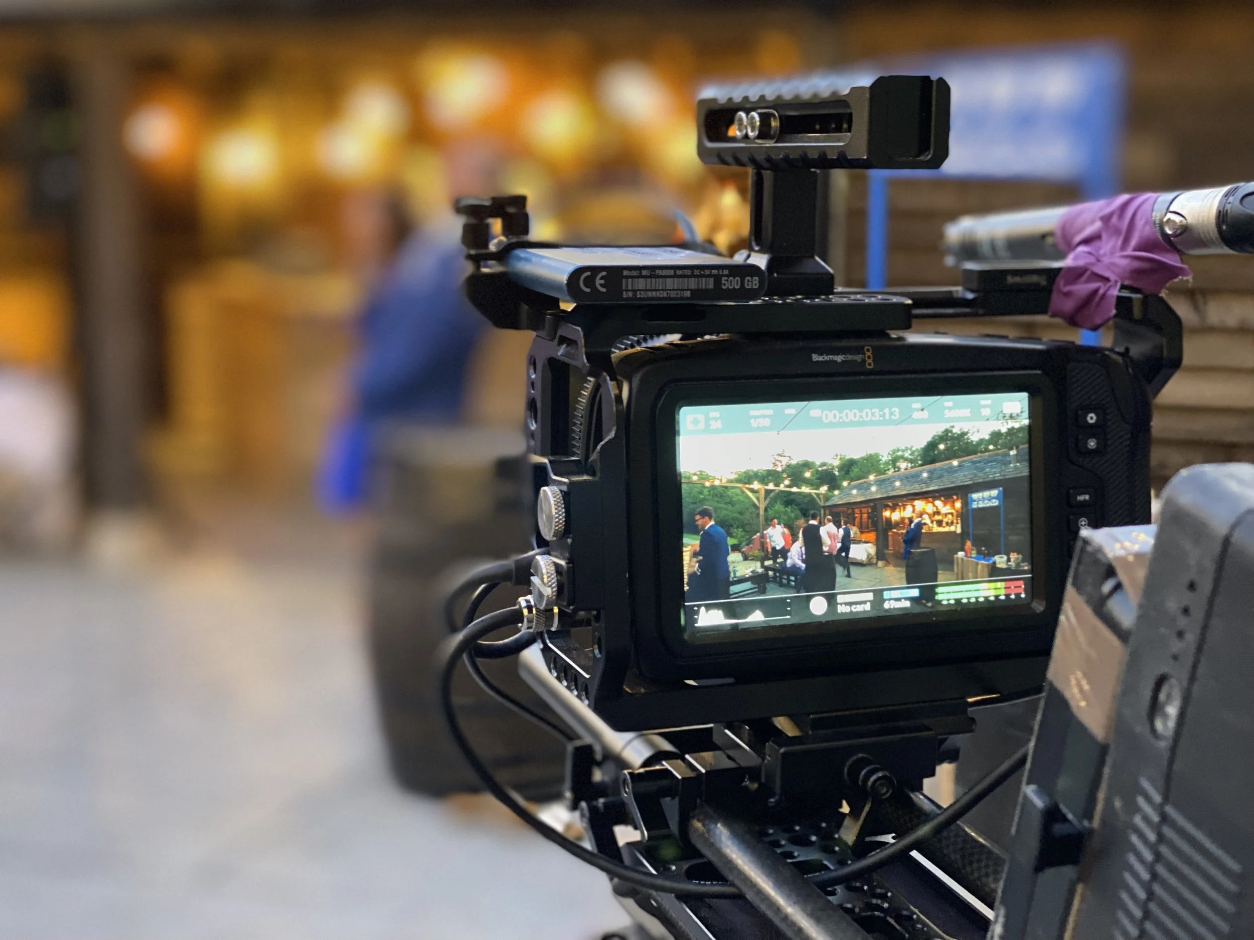 A professional camera on a film set captures a scene of people outdoors at a market or gathering during evening time, with a blurred background of people and lights.