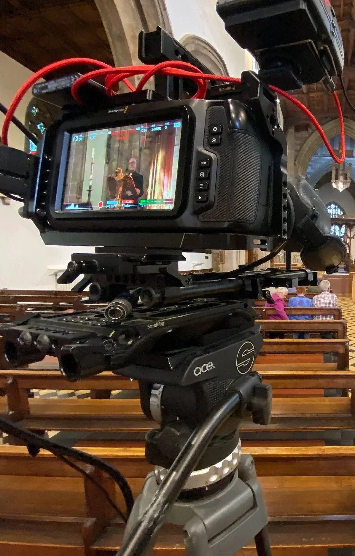 A professional video camera on a tripod recording inside a church, with a few seated people visible in the background.