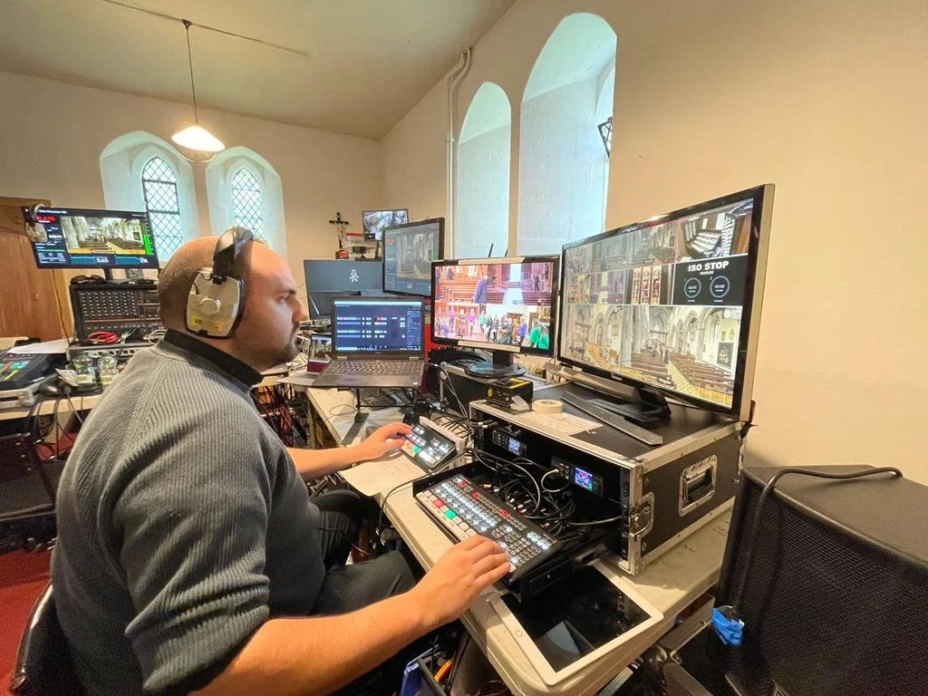 Man in gray sweater wearing headphones operating video production equipment with multiple monitors and control panels in a room with arched windows.