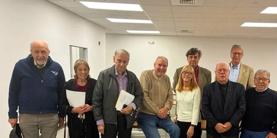 Group of nine older adults standing in a room with white walls and ceiling tiles, some holding papers, smiling at the camera.