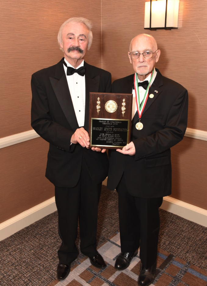 Two elderly men in tuxedos standing in a room, holding a plaque, with one wearing a medal around his neck.