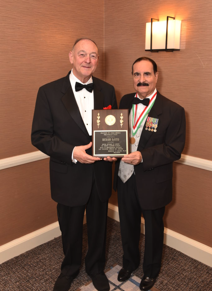 Two men in tuxedos standing, holding a plaque and medal, at an awards ceremony. One man has a bald head and the other has dark hair and a mustache,双方 smiling. Background has a brown wall and a wall light.