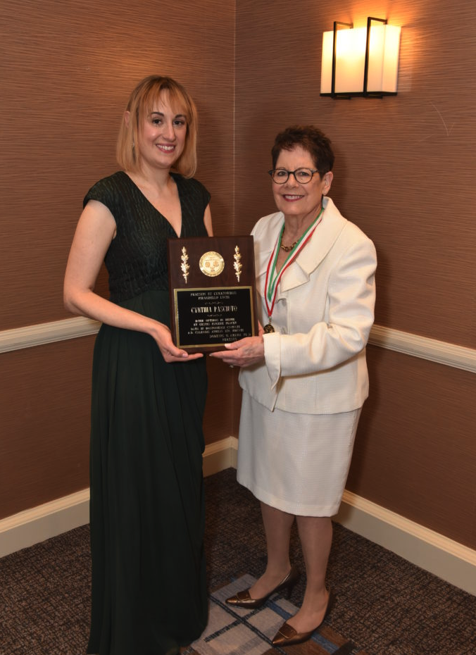 Two women standing in a room with brown walls, smiling, holding a plaque and a medal, celebrating an award moment.