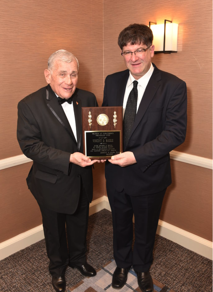 Two men in formal suits holding an award plaque, standing in a room with brown walls and a wall-mounted light fixture.