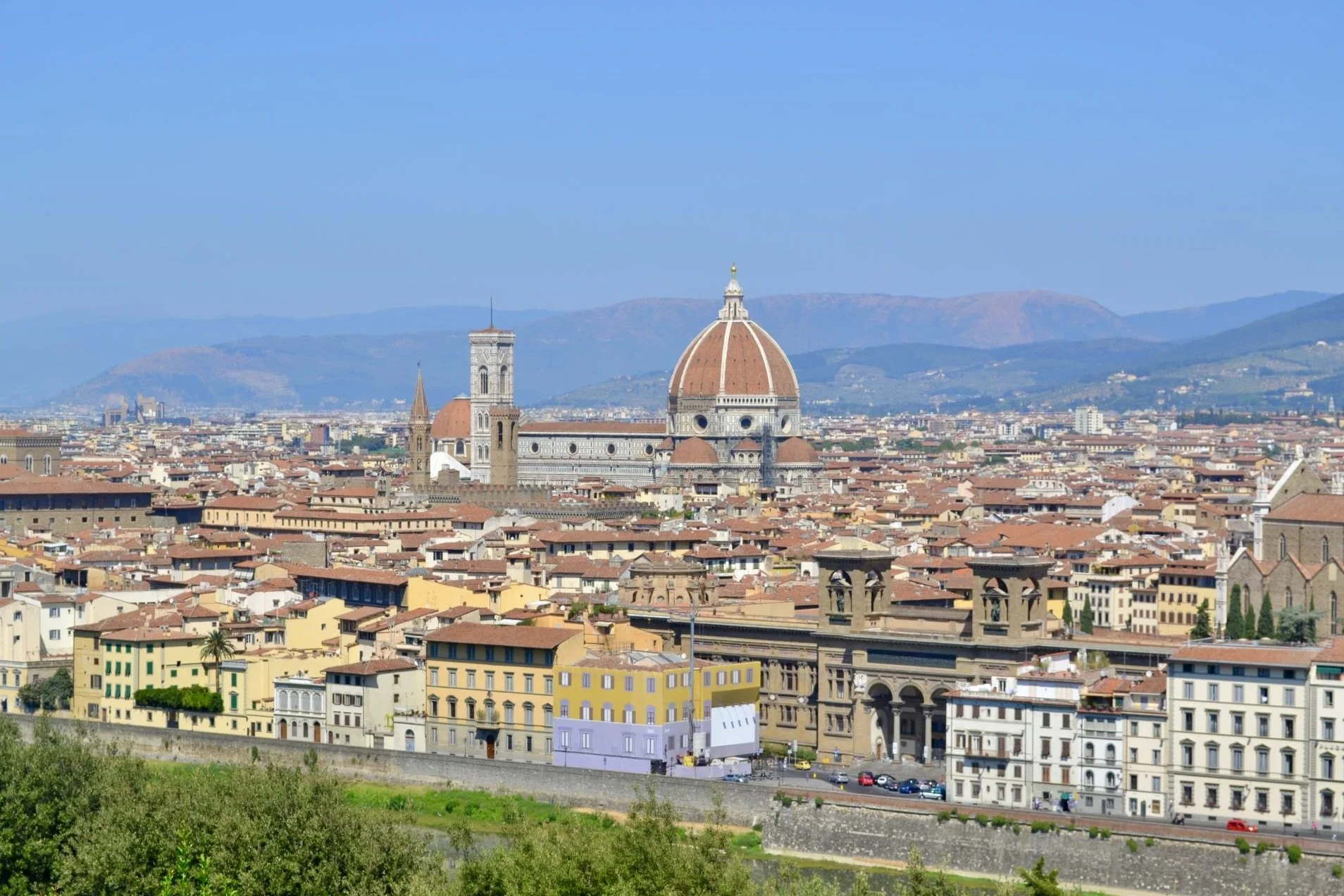 Panoramic view of Florence, Italy, featuring the Florence Cathedral with its prominent red-tiled dome and a cityscape of historic buildings, with mountains in the background.
