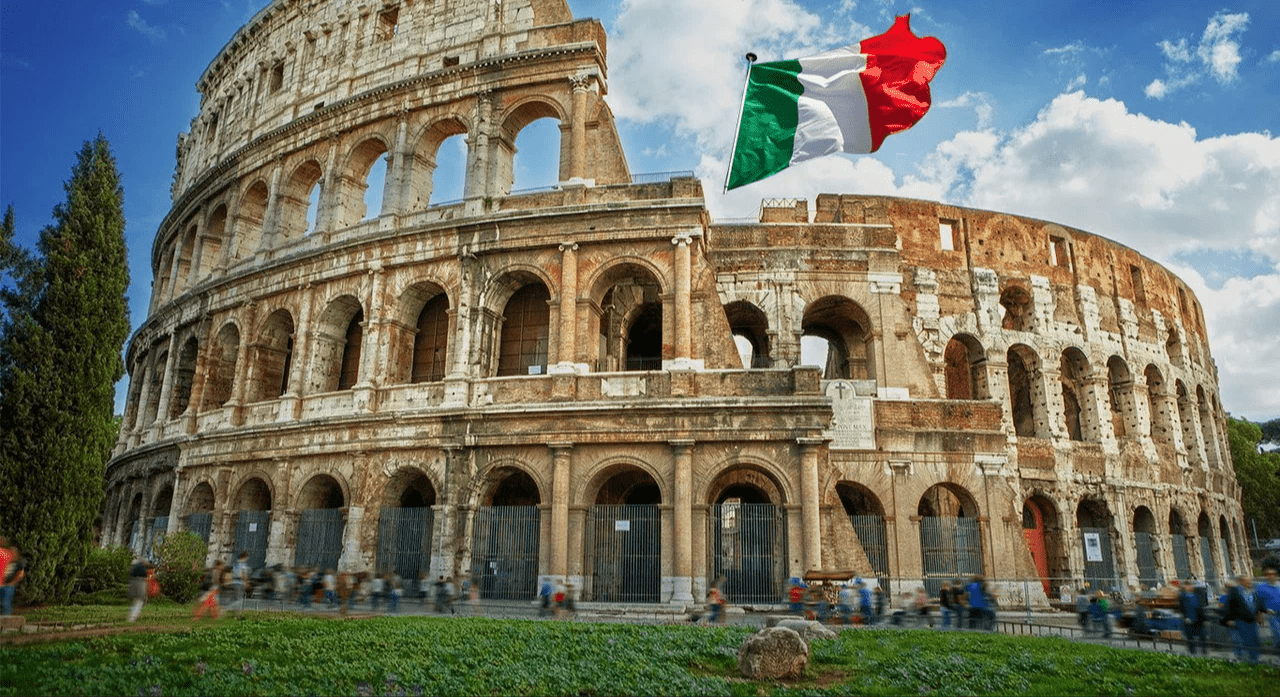 The Colosseum in Rome, Italy, with an Italian flag flying in the sky nearby, and people walking in front of it on a sunny day.