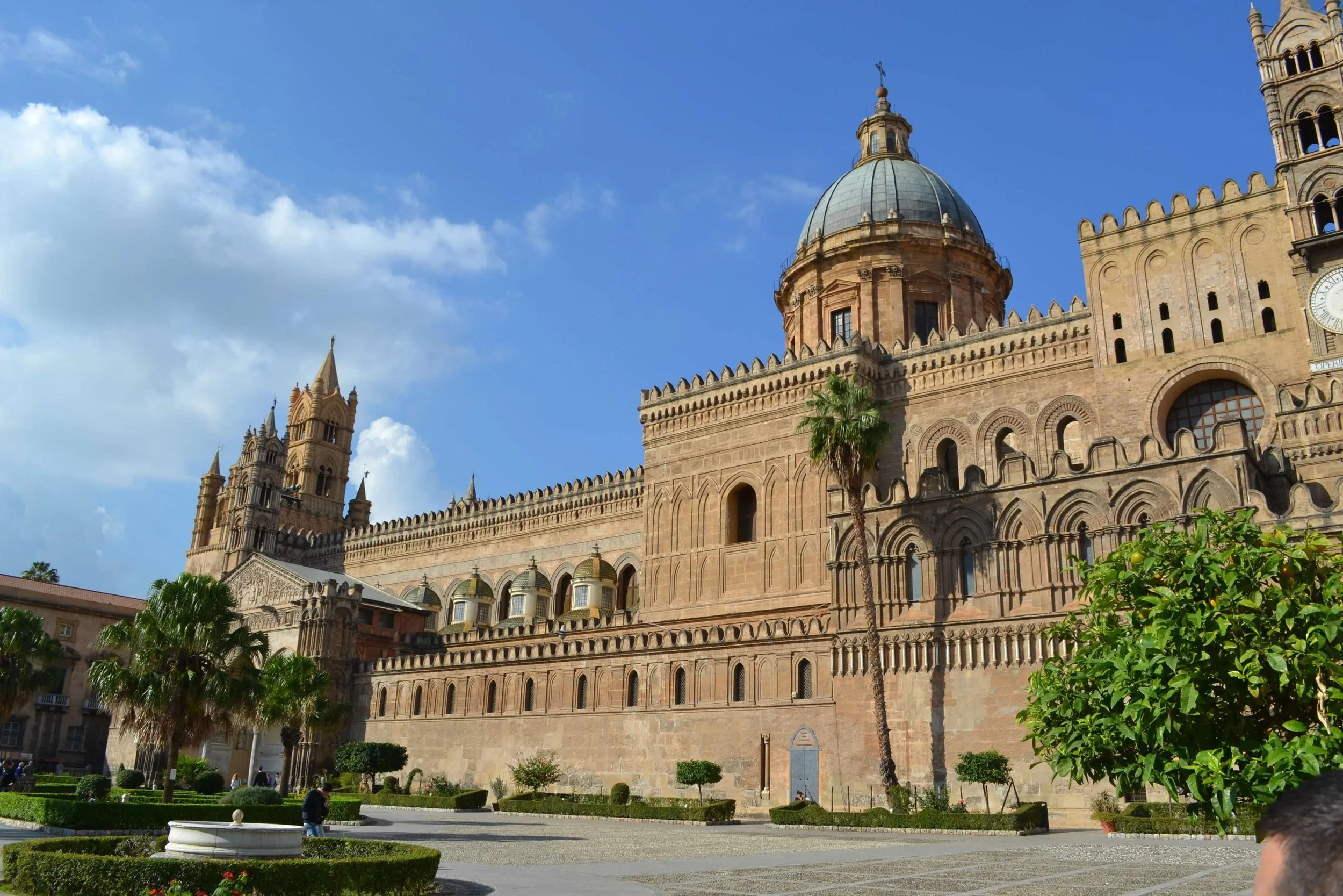 Facade of a large historic cathedral with a domed roof and tall spires, surrounded by palm trees and greenery, under a blue sky with some clouds.