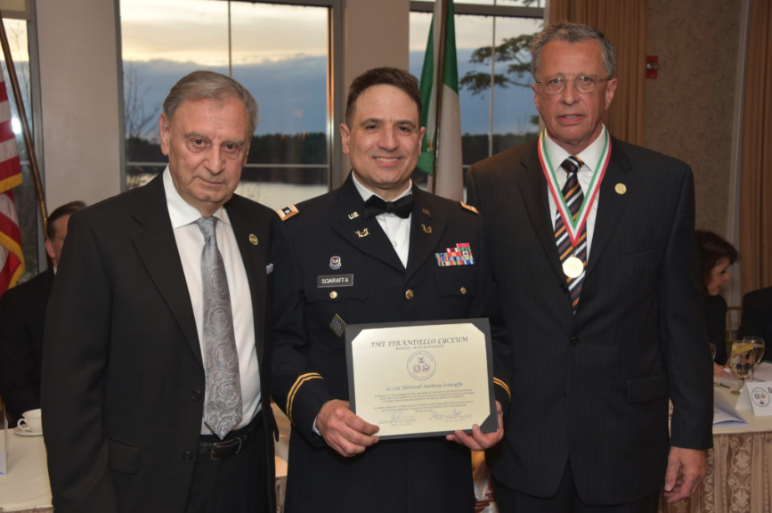 Three men in formal attire standing together at an award ceremony, with the man in the middle holding a certificate.