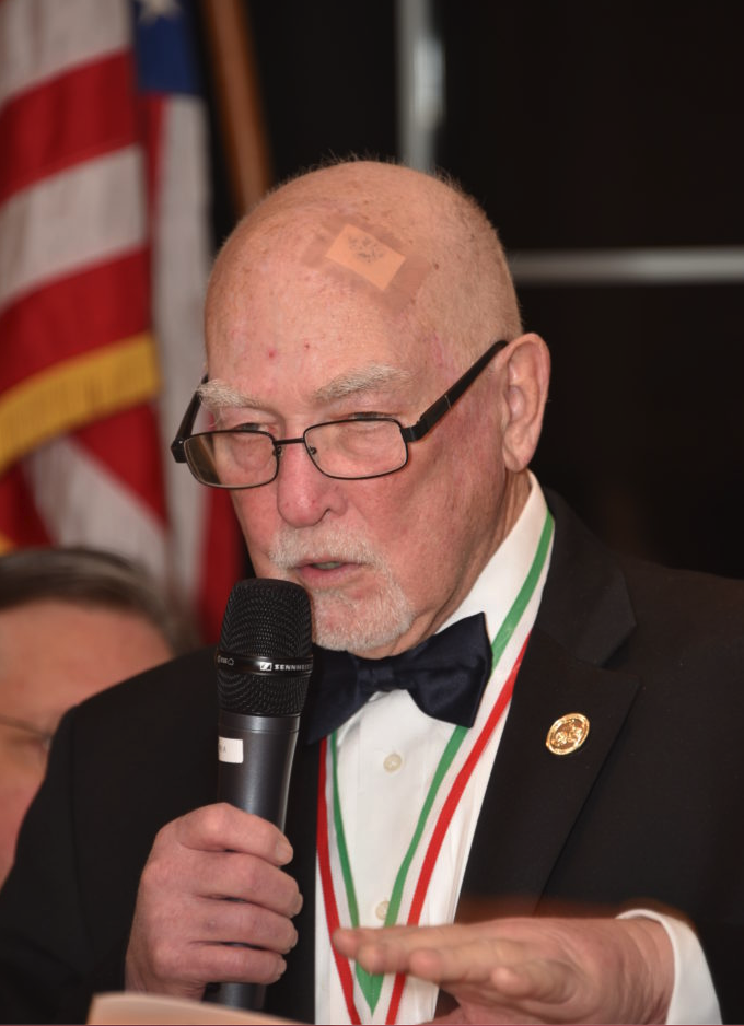 An elderly man in a tuxedo with a bow tie, glasses, and a gold lapel pin is speaking into a microphone at a formal event. He is wearing medals around his neck and has a bandage on his forehead. An American flag is visible in the background.