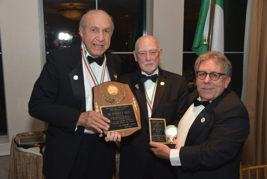 Three men in tuxedos at an awards ceremony, with one holding a plaque and the other holding a small trophy.