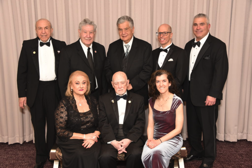 Eight formal-dressed individuals posing for a photo in front of beige curtains at an event.