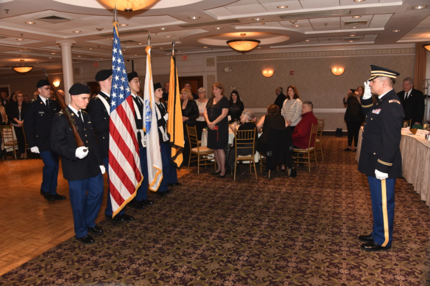 Military Color Guard presenting flags in a dining or banquet hall with attendees observing.