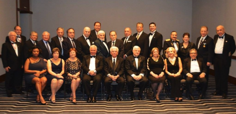 A group of well-dressed men and women in formal attire posing for a group photo in a room with a plain wall and striped carpet.
