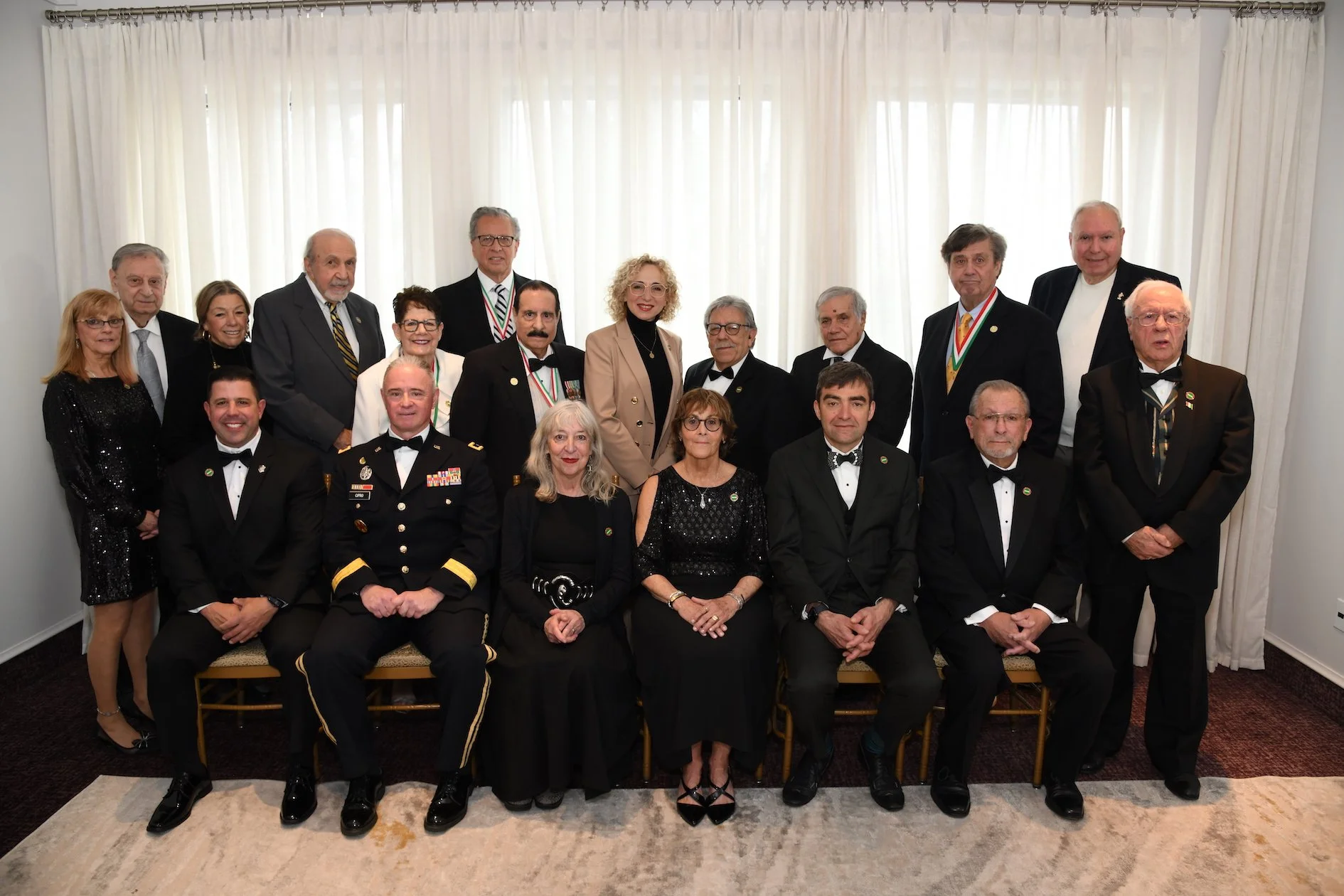Group of people, mostly men in formal tuxedos, some with medals or ribbons, posing for a photo in a room with white curtains.
