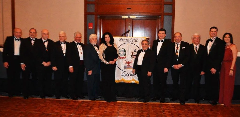 Group of people in formal attire posing for a photo at an event, with a banner reading 'Pirandello Lyceum' in the background.