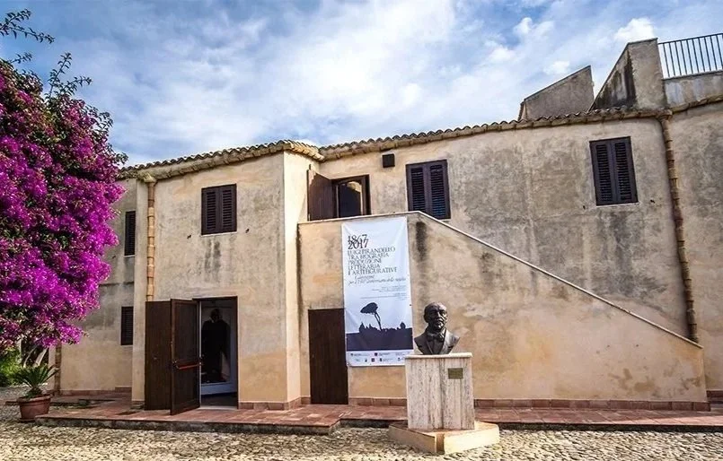 A historic building with beige stucco walls, dark wooden shutters, and a staircase on the right side. There is a bust statue on a pedestal in front, and a purple flowering tree on the left. A poster referencing 1867 hangs on the building, with a partly cloudy sky overhead.